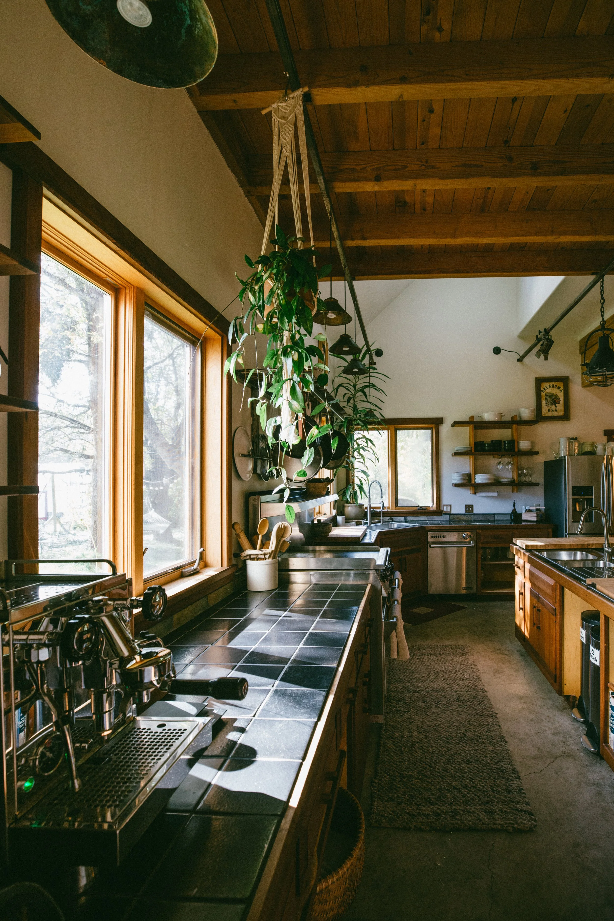A cozy kitchen with wooden cabinets and black tile countertops, large windows allowing natural light, hanging plants, and various kitchen utensils and appliances visible.