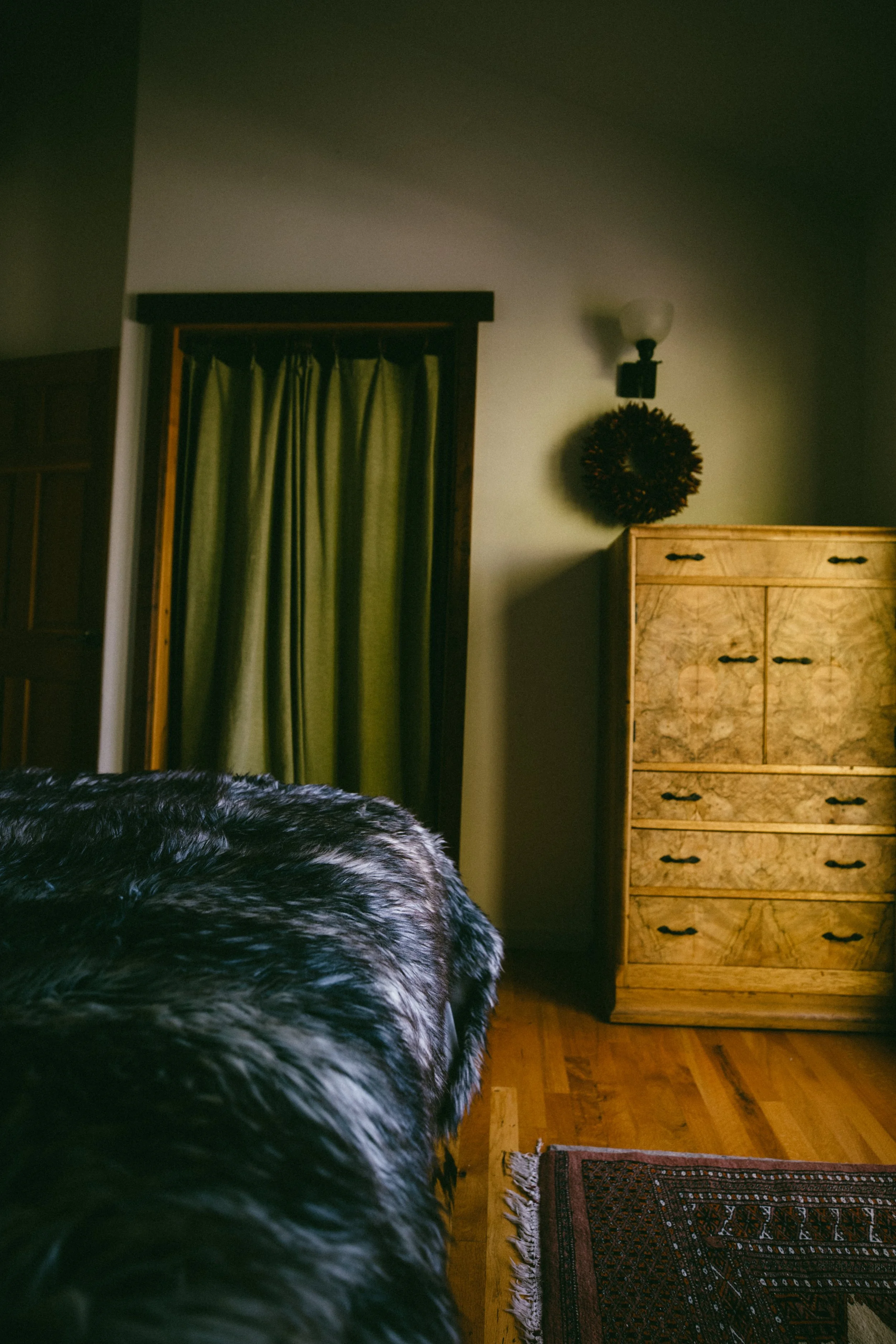Part of a bedroom with a black and white fur blanket, a wooden dresser, a wall-mounted light fixture, a decorative wreath, green curtains, and a partial view of a wooden door and ceiling.