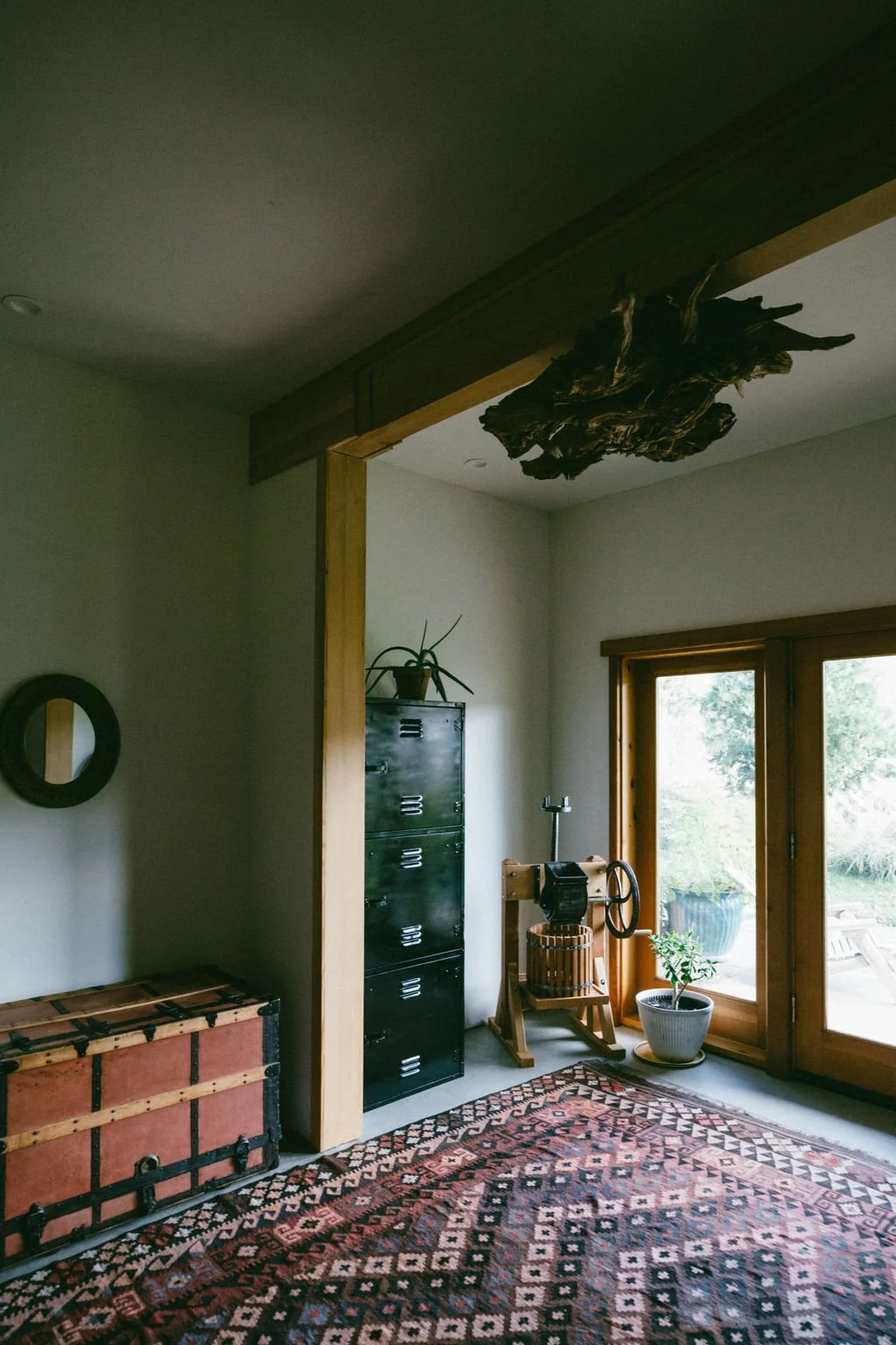 Interior of a room with a large window, potted plants, a black metal locker, an antique chest, and a wooden device with a wheel, with a patterned rug on the floor.