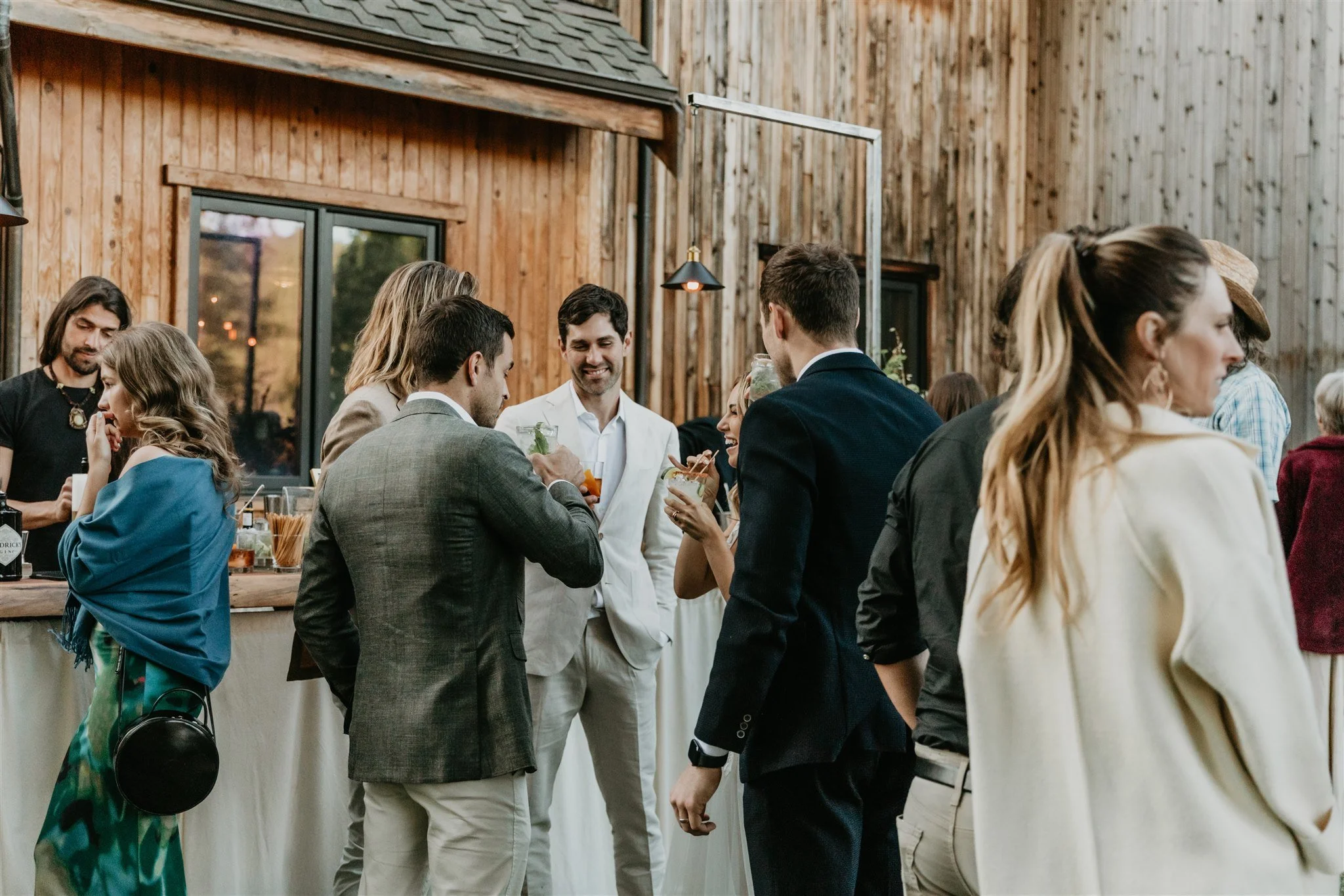 Group of people socializing at a bar or reception area with wooden walls, some holding drinks, engaged in conversations.