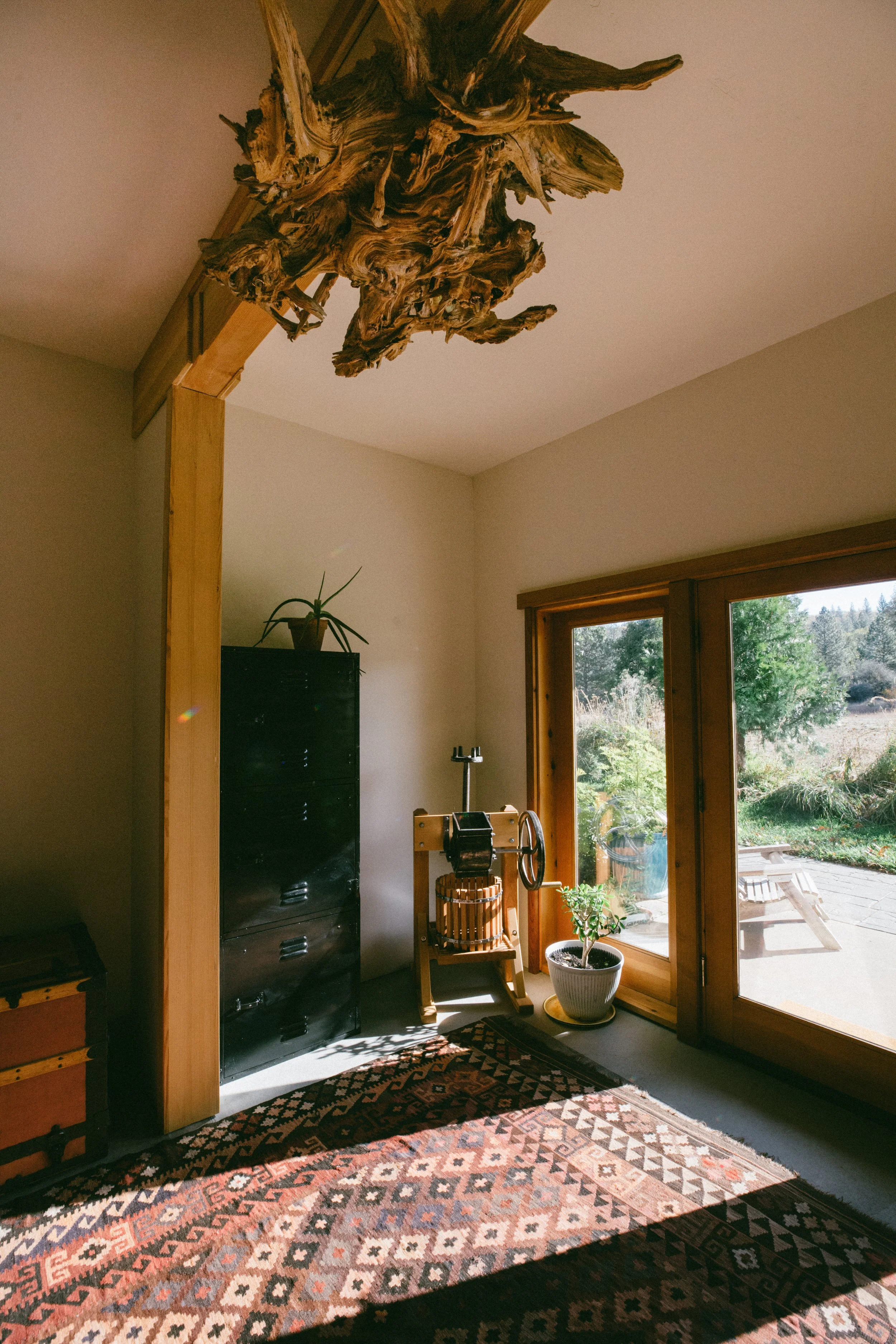 A cozy room with a wooden beam, natural sunlight, a black storage cabinet, a potted plant, a small water wheel, an outdoor seating area visible through glass doors, and a patterned rug.