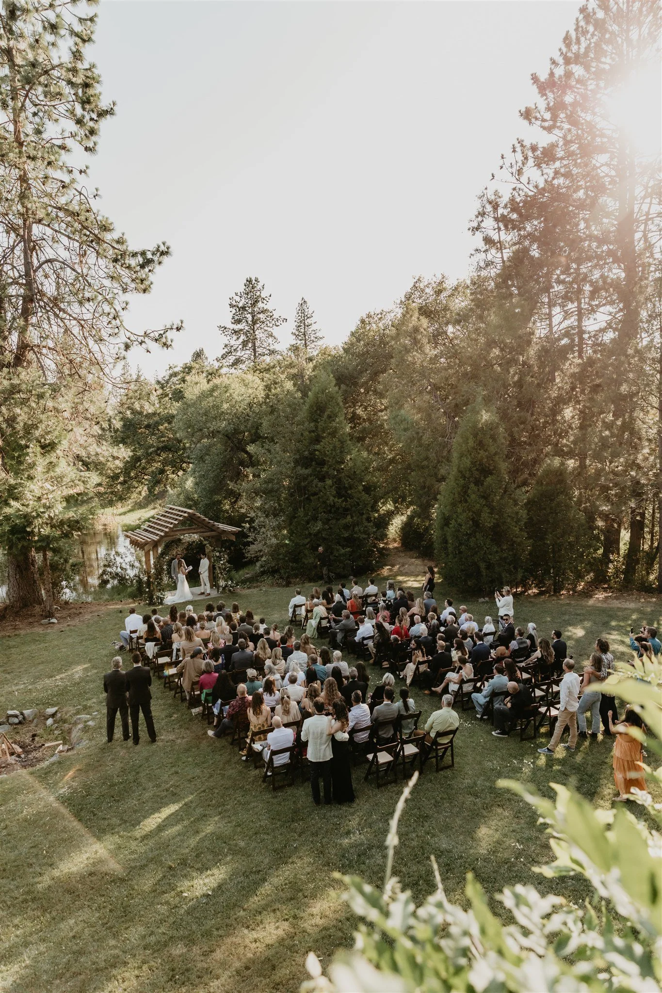 Outdoor wedding ceremony with a large congregation of guests seated on chairs in a grassy area surrounded by tall trees, with two people standing at the altar.