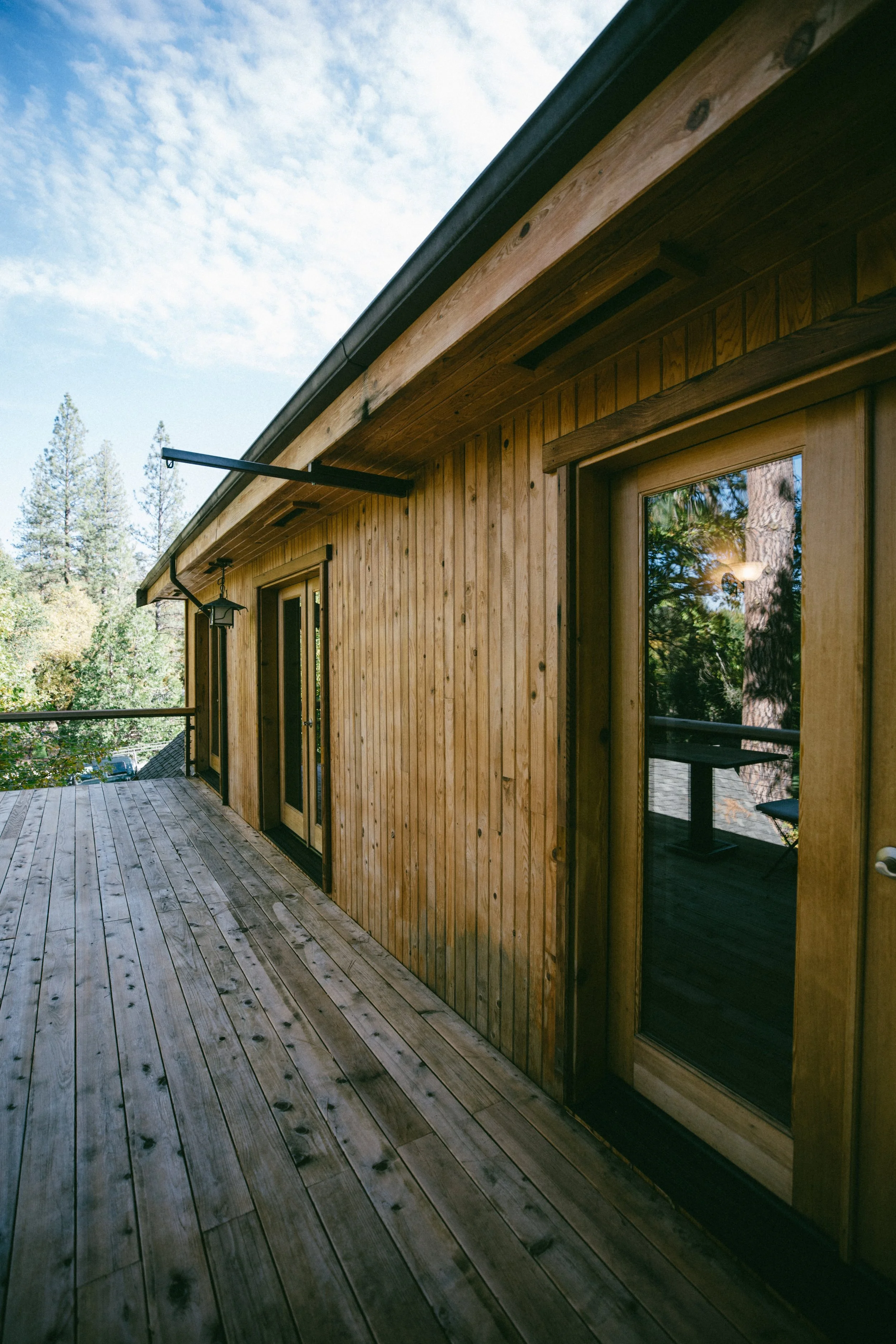 Wooden exterior balcony with sliding glass doors and a railing, surrounded by tall trees and a partly cloudy sky.