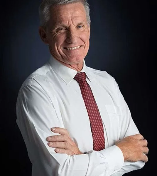 A smiling older man with gray hair, wearing a white dress shirt and a red tie, standing with arms crossed against a dark background.