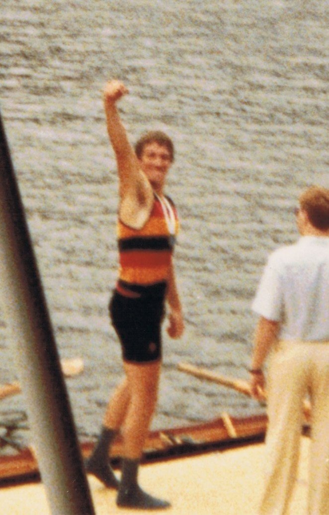 A man in a sleeveless striped athletic shirt and shorts is raising his arm in front of a brick wall. A person in a white shirt and beige pants is standing nearby, holding a stick.