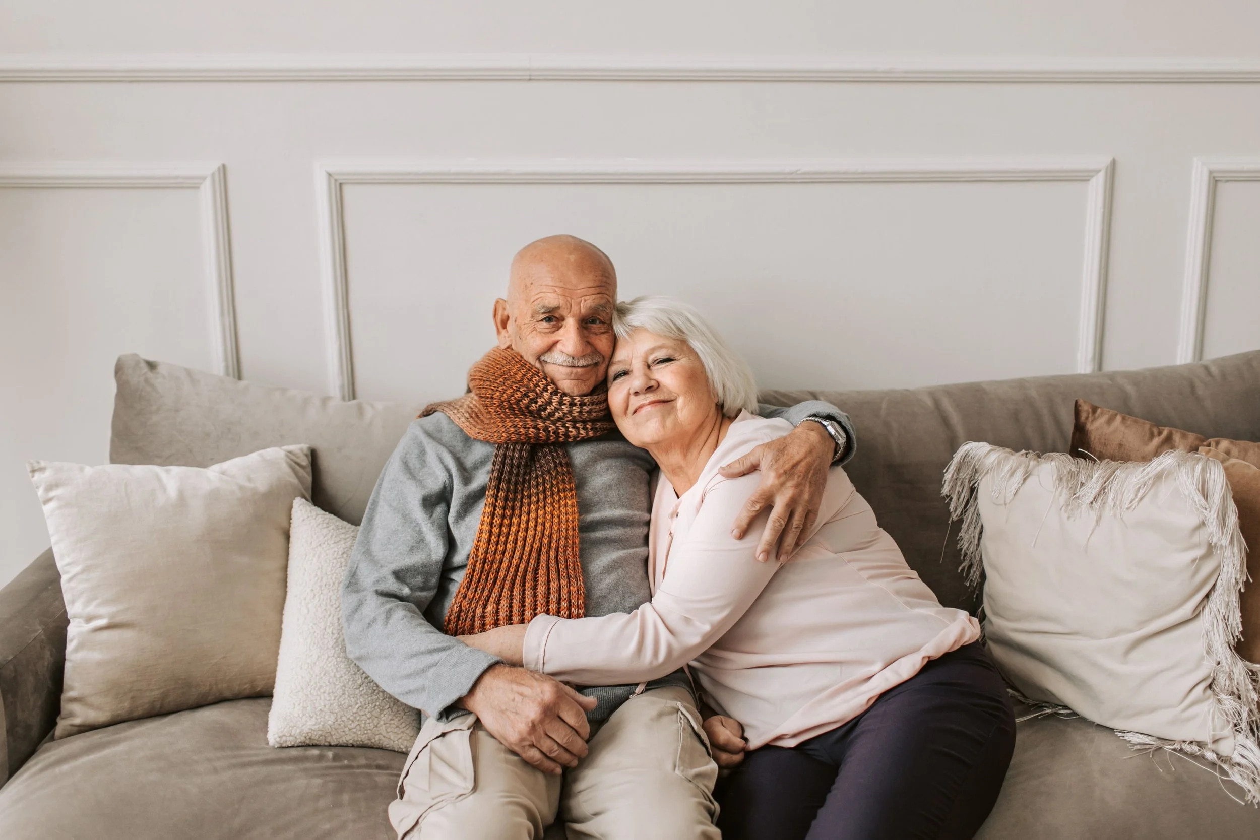 An elderly couple hugging and smiling on a beige couch in a living room.