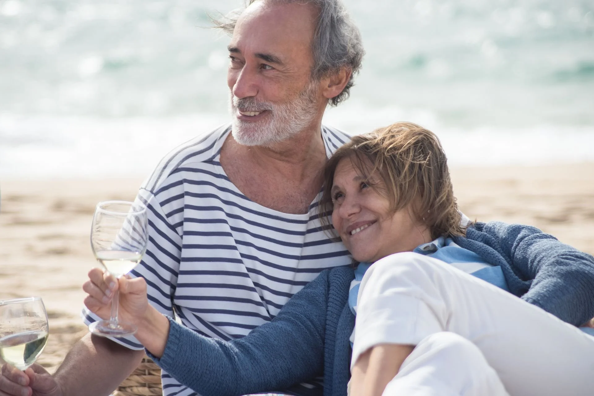 Two older adults, a man and a woman, sharing a joyful moment on the beach, with the man holding a glass of white wine and both smiling amid the ocean background.