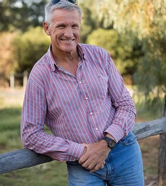 Smiling older man in a pink striped shirt and jeans leaning on a wooden fence outdoors with trees in the background.