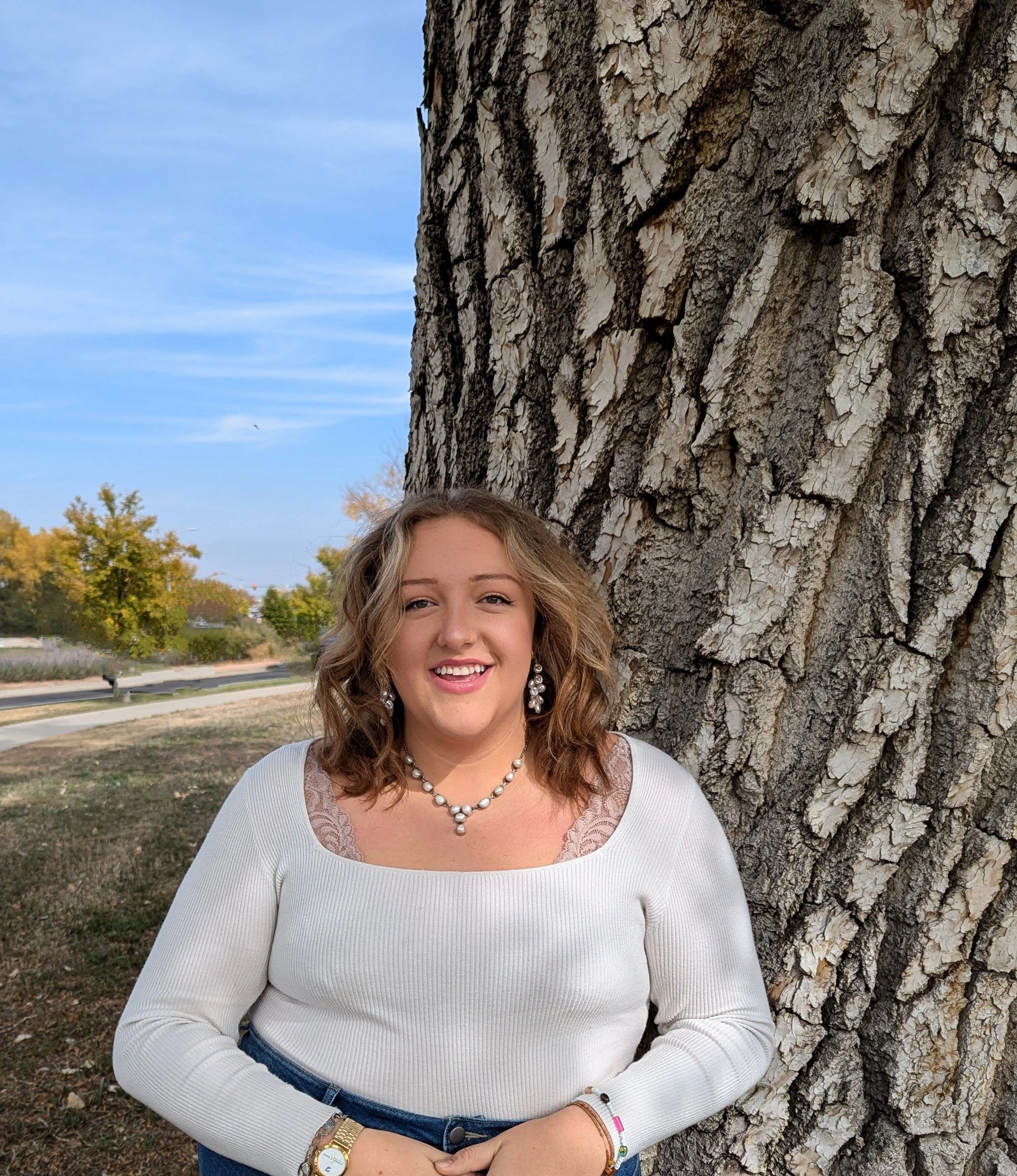 A woman with wavy blonde hair smiling, wearing a white long-sleeve top with lace details, jewelry including a pearl necklace, earrings, and a bracelet, standing outdoors near a large tree with rough bark. The background shows a partly cloudy blue sky, trees with fall foliage, and a park bench.