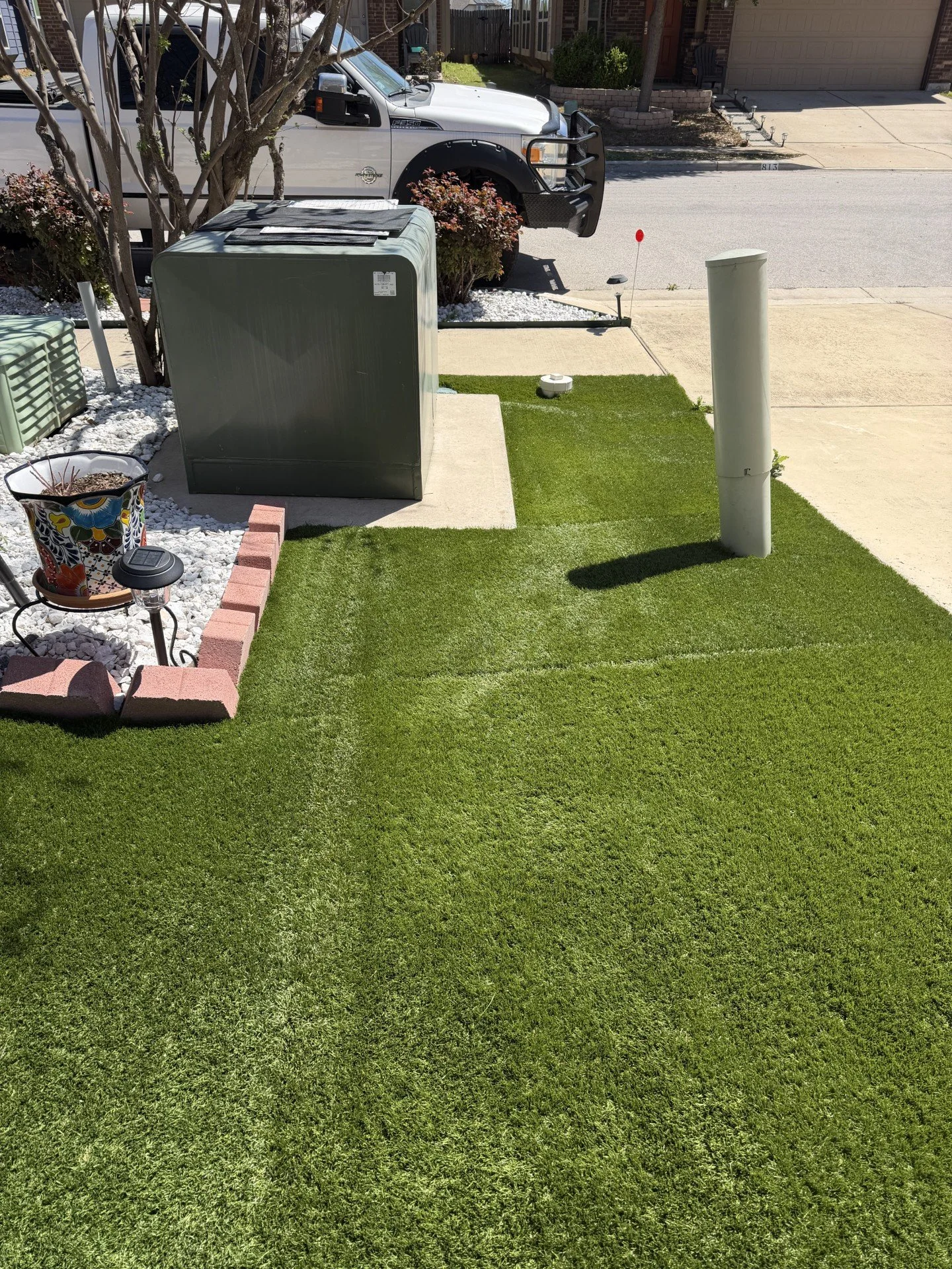 A well-maintained front yard with green grass, a small brick border garden bed, a decorative flower pot, a lamp, a large gray utility box, a white pipe, a sidewalk, and a street with a white truck parked in front of a house.