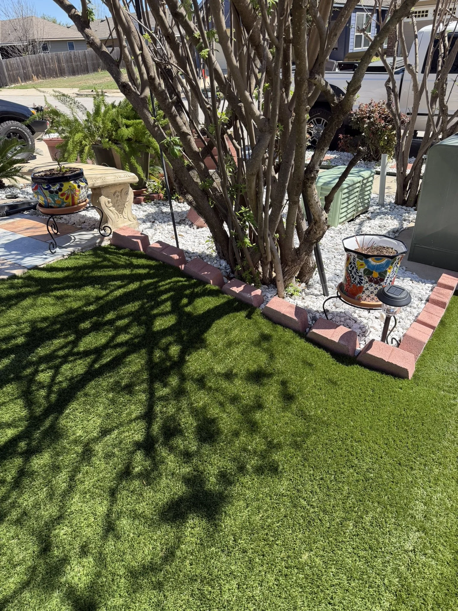 A well-maintained backyard with a green lawn, a large tree casting shadows, colorful flower pots, white gravel, and some outdoor furniture and decoration.