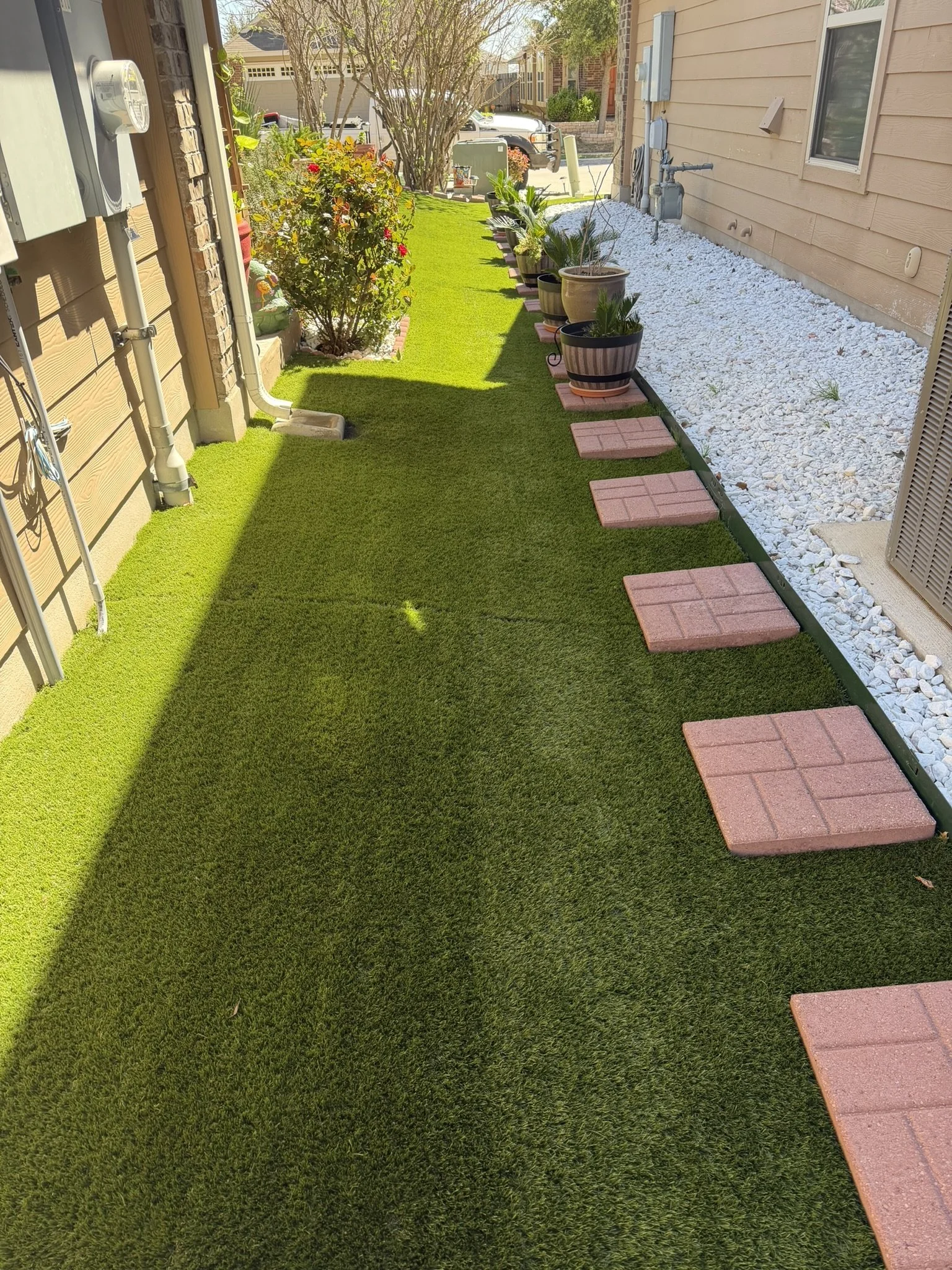 A backyard patio with artificial grass, potted plants, and stepping stones, bordered by white rocks, with a house wall on one side and a yard with trees and vehicles in the background.