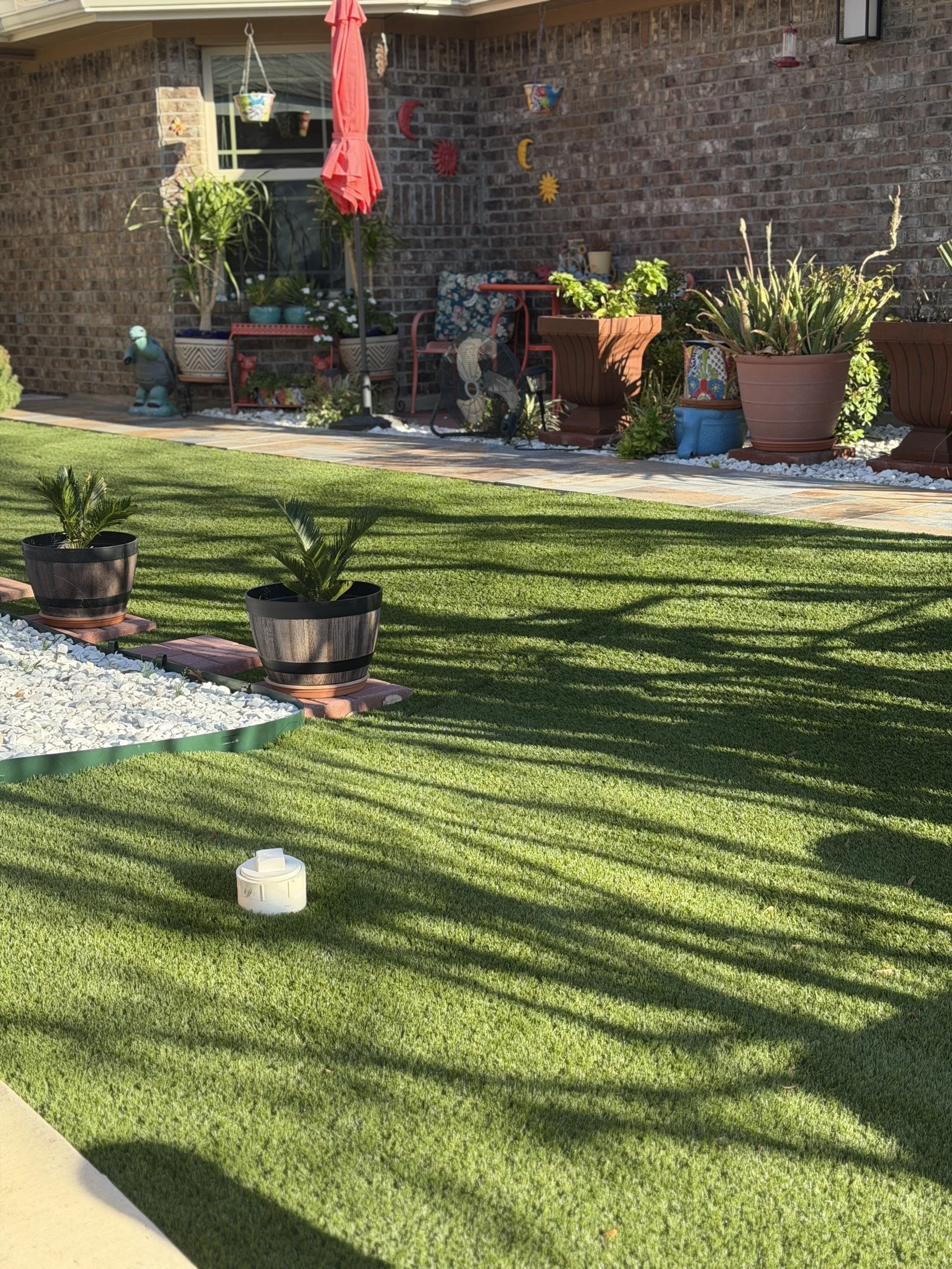 Backyard garden with potted plants, lawn, outdoor furniture, and decorative items against a brick wall.