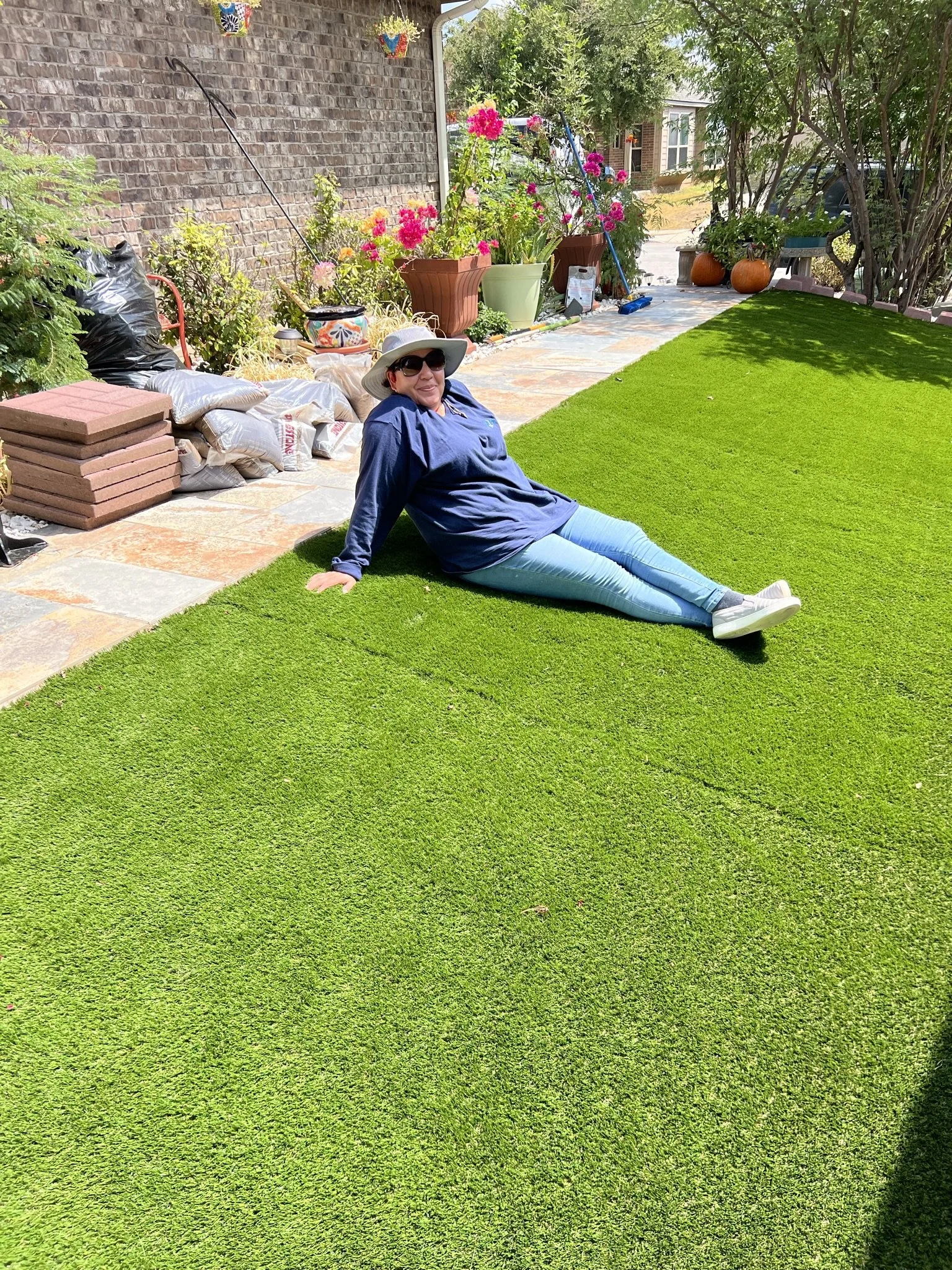 Woman sitting on a vibrant green lawn in a garden, wearing sunglasses, a wide-brimmed hat, a blue shirt, and light blue jeans, smiling at the camera.