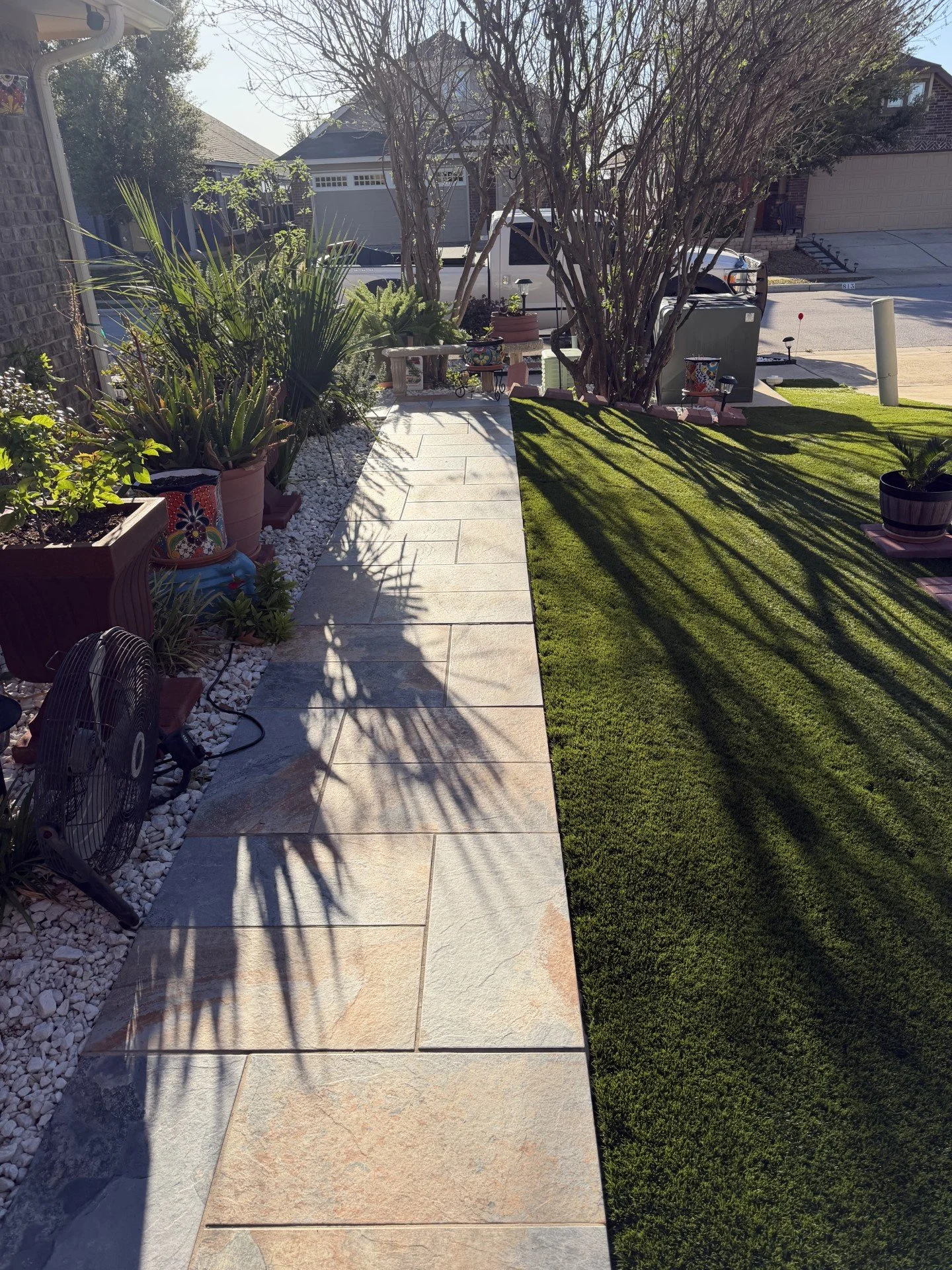 A backyard with a stone pathway on the left, green lawn on the right, potted plants along the pathway, a large tree casting shadows, and a street with a white truck and houses in the background.