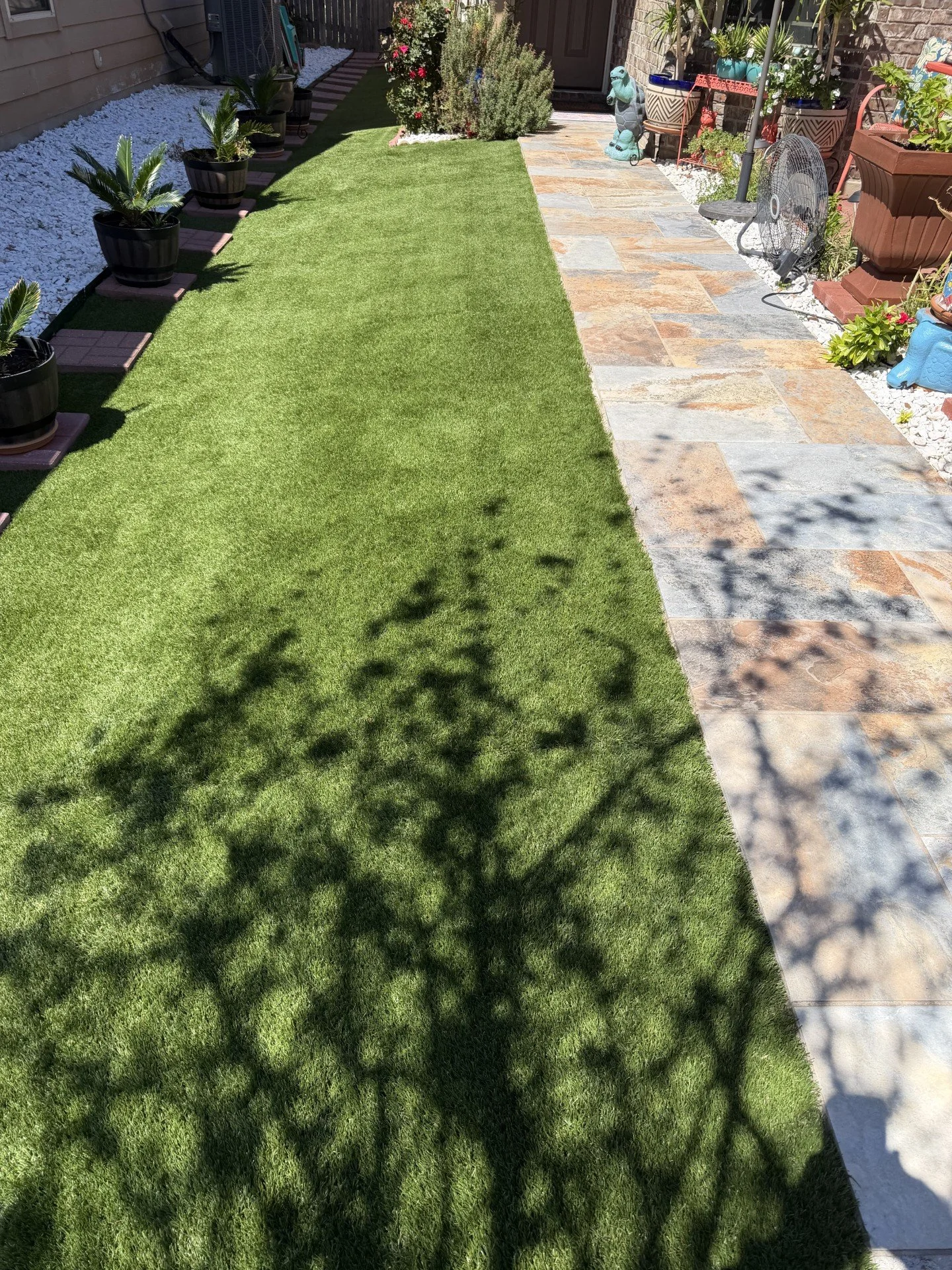 A well-maintained backyard with a strip of green artificial grass, potted plants along the left side, and a tiled patio with garden ornaments and a fan on the right side.