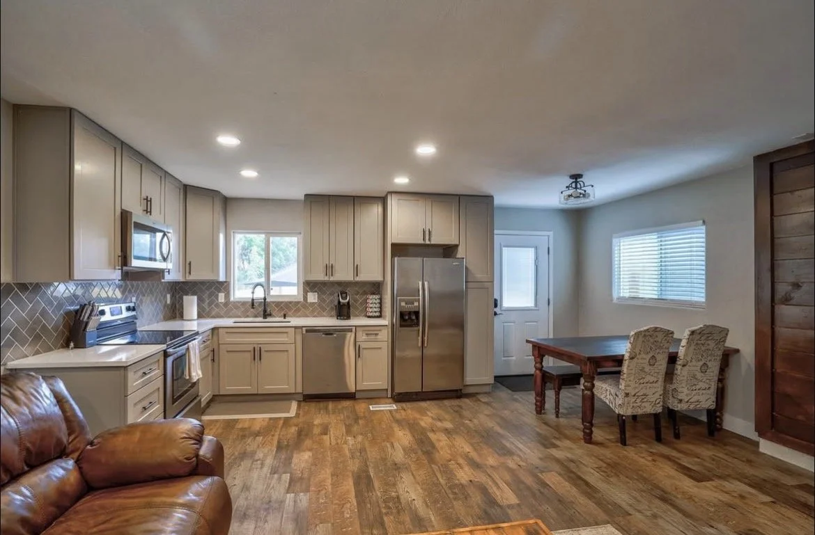 Open concept kitchen with gray cabinets, stainless steel appliances, a small dining area with a wooden table and two patterned chairs, hardwood floors, and a window and door with blinds.