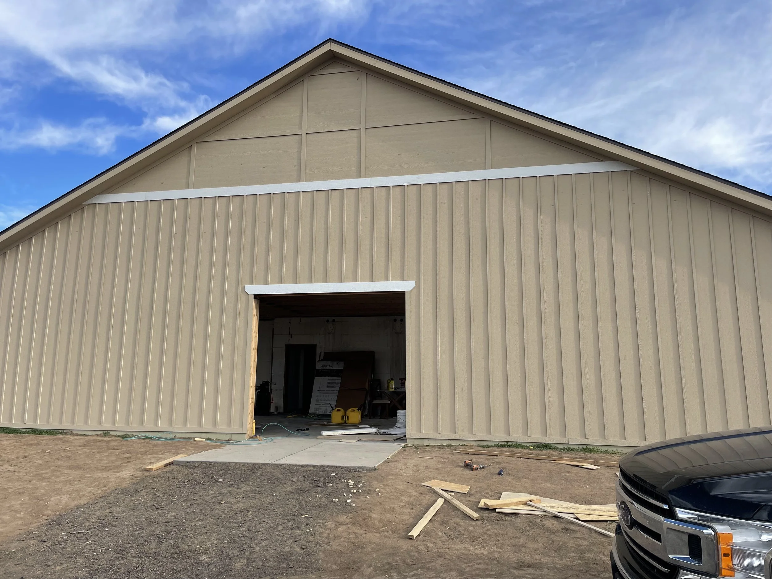 A building under construction with beige metal siding, an open garage door, and construction tools inside. The sky is partly cloudy with blue sky and clouds visible. There are construction materials on the ground and part of a black vehicle is seen o