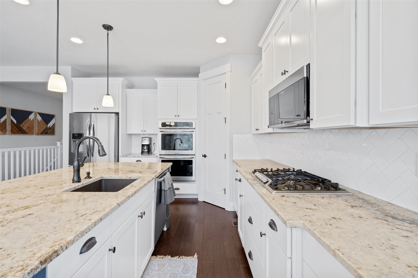 Modern kitchen with white cabinetry, granite countertops, stainless steel appliances, a double oven, a microwave, a gas stove, a coffee machine, and a sink with a pull-down faucet. Pendant lights hang over the island, and a dark wood floor runs throu