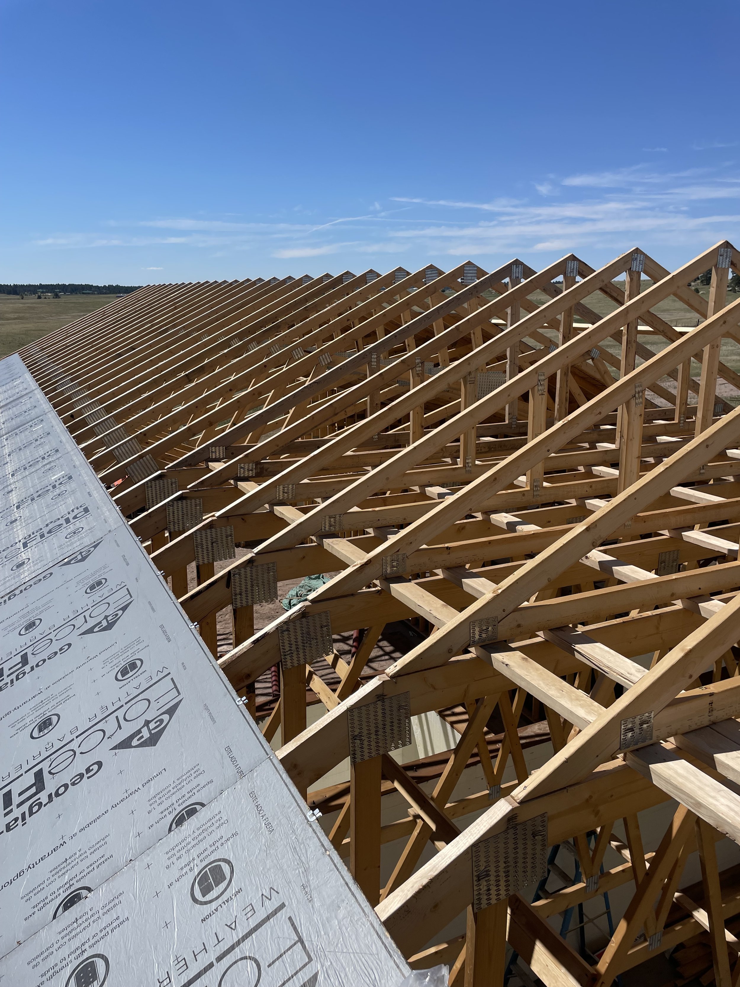 Construction of a roof with wooden trusses and underlayment, set against a blue sky and open landscape