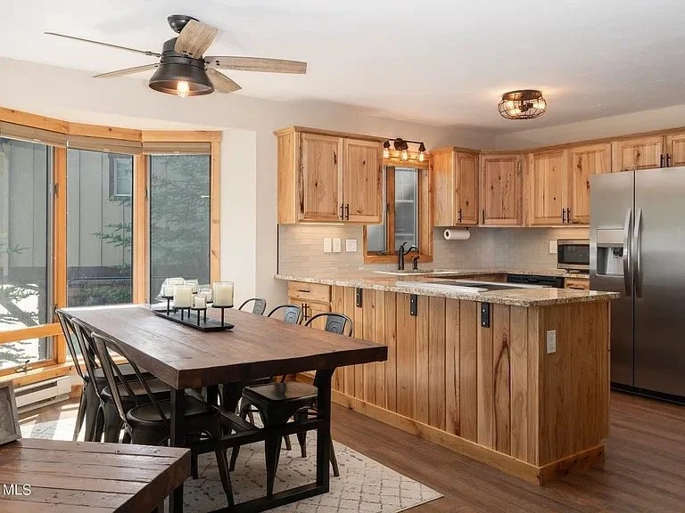 Condo kitchen and dining area with wooden cabinetry and a large wooden dining table with chairs, large windows, and stainless steel appliances.