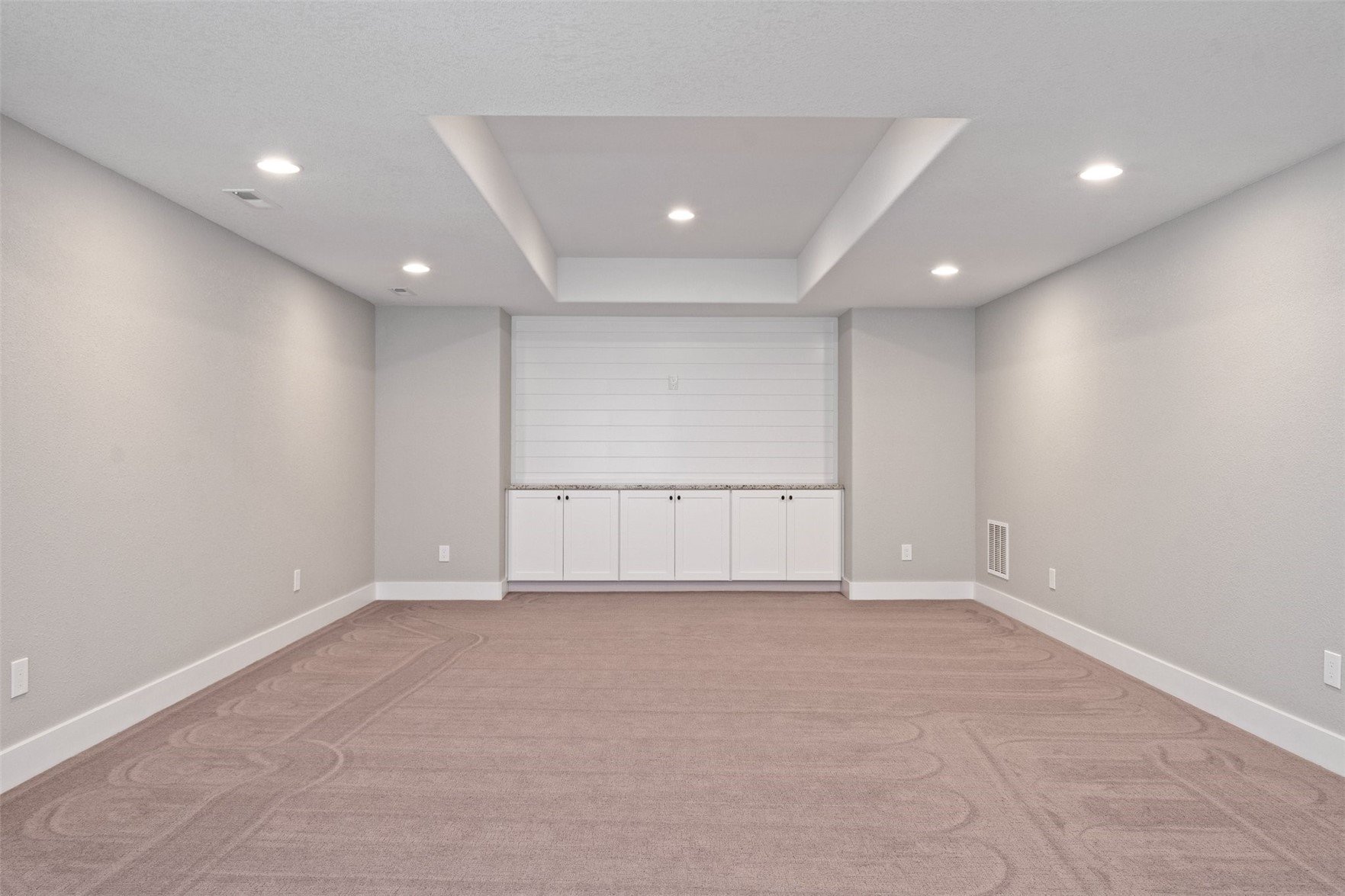 Empty basement room with carpet, white walls, recessed ceiling lights, built-in white cabinets, and a large window with blinds.