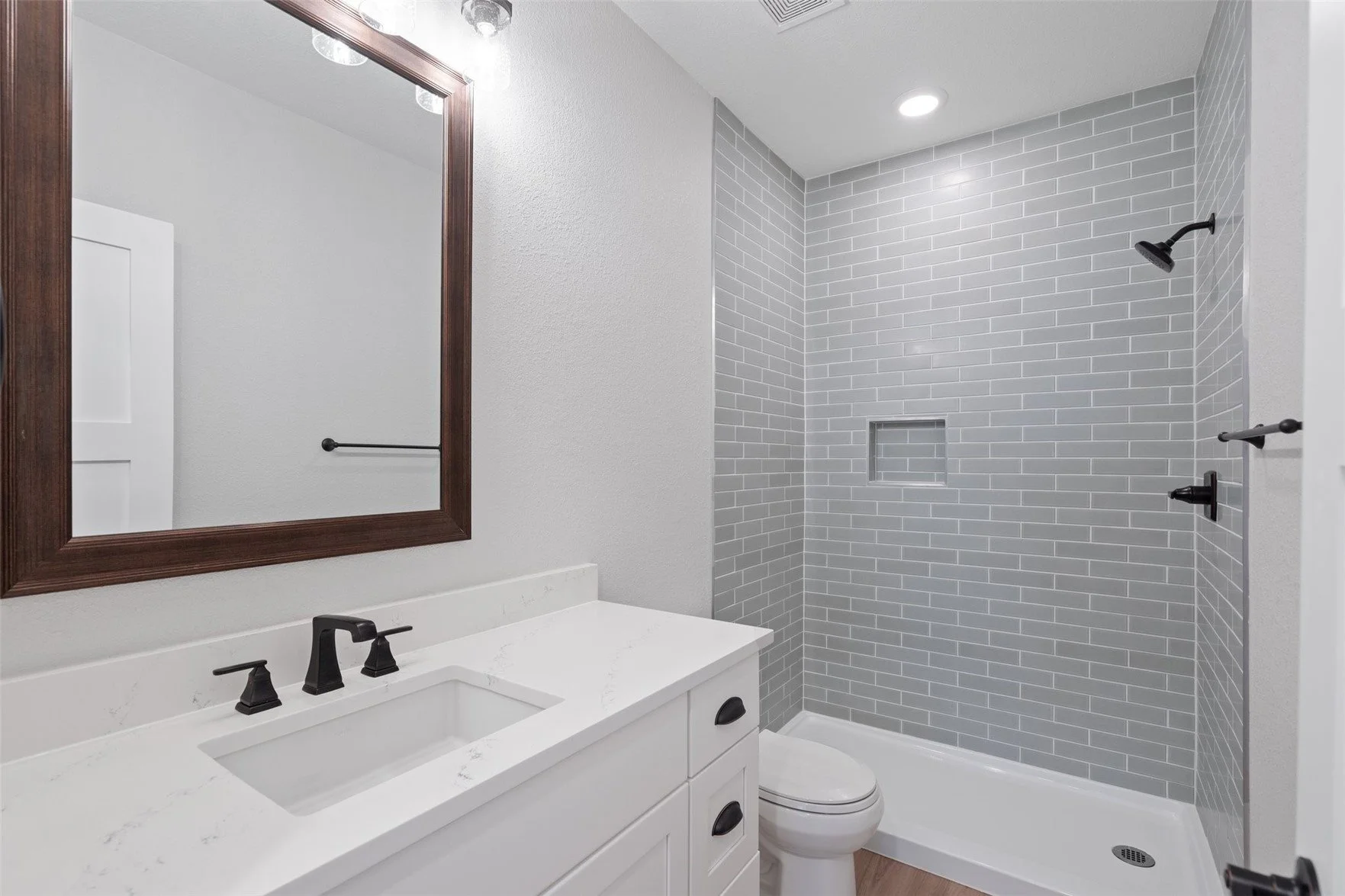 Modern bathroom with white vanity, black faucet, large mirror, toilet, and walk-in shower with gray subway tile walls and built-in niche.