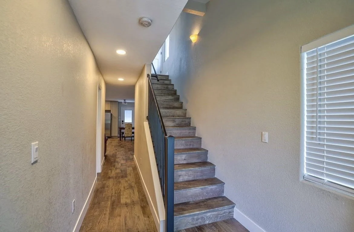 Interior hallway with wooden flooring, beige walls, and a staircase leading upstairs on the right side.