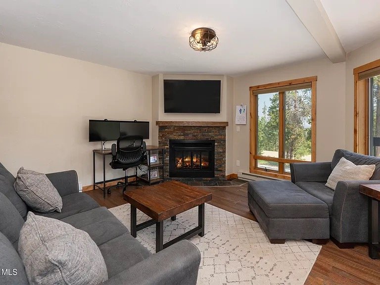 Living room with a fireplace, a mounted television above it, a window showing trees outside, and seating including a gray sofa and an armchair. There's a small desk with a computer and a chair, and a wooden coffee table in the center.