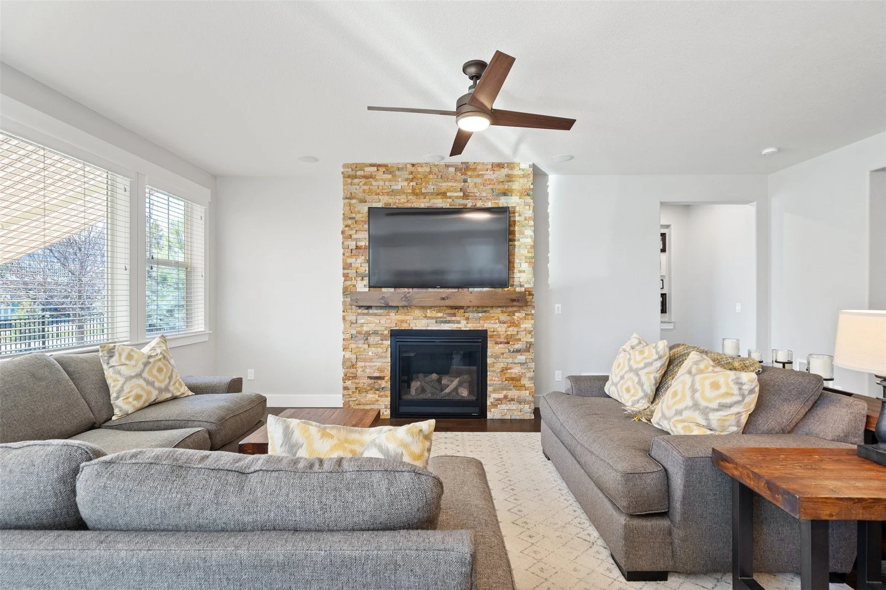 Living room with a central stone fireplace, mounted flat-screen TV above the fireplace, ceiling fan, large windows on the left, gray sofas with patterned pillows, wooden side table with lamp, and a beige area rug.