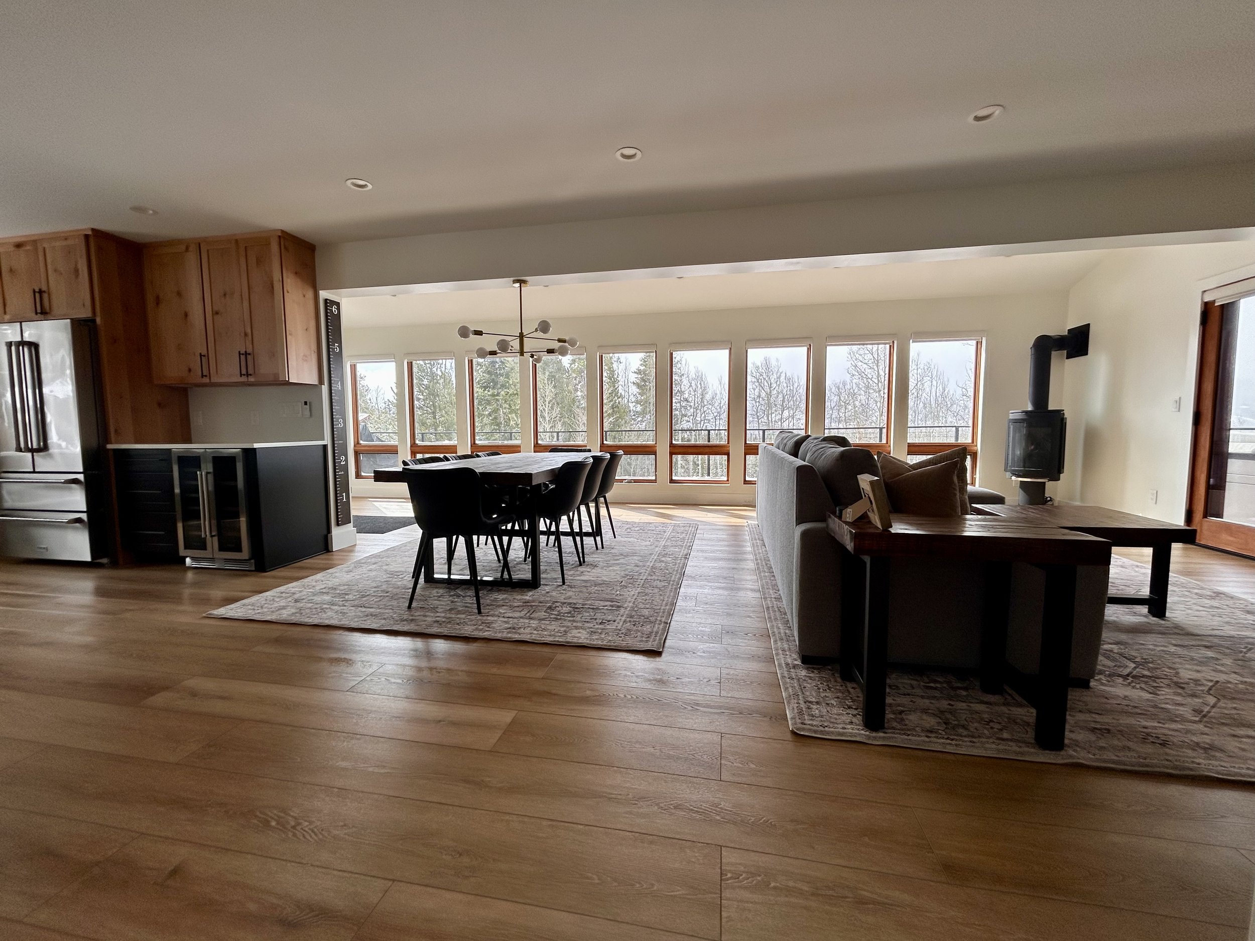 Open-concept living and dining area with large windows, wooden flooring, a modern chandelier, a dining table with chairs, a gray sofa, a coffee table, a wood stove, and a sliding glass door.