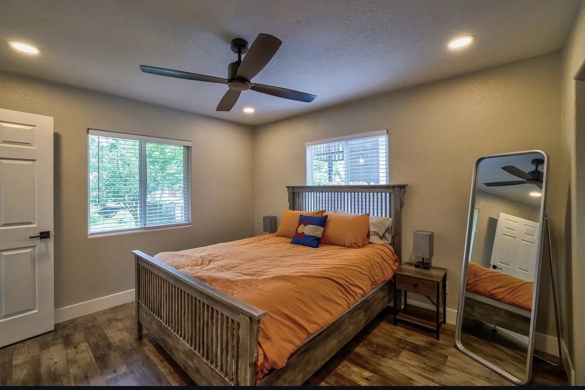 A bedroom with a wooden bed frame and orange bedding, a small wooden nightstand, a full-length mirror, two windows with blinds, and a ceiling fan.