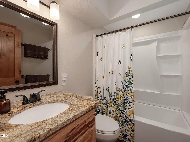 Bathroom with wooden cabinet, granite countertop, sink, mirror, white shower with floral curtain, small towels, and ceiling lighting.