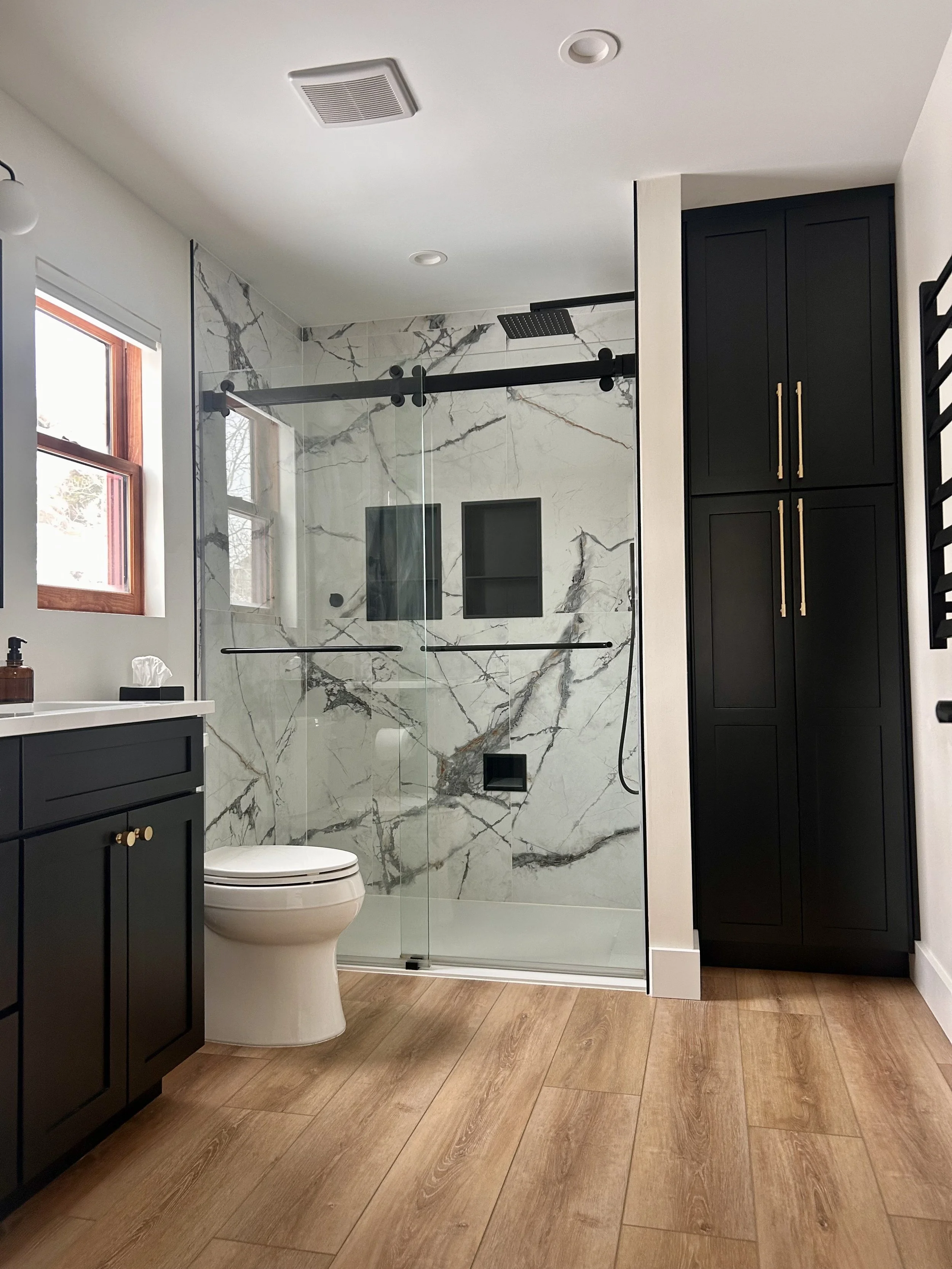 Modern mountain bathroom with white walls and wooden floors, black cabinetry, and a marble-tiled walk-in shower with black fixtures and two niches.