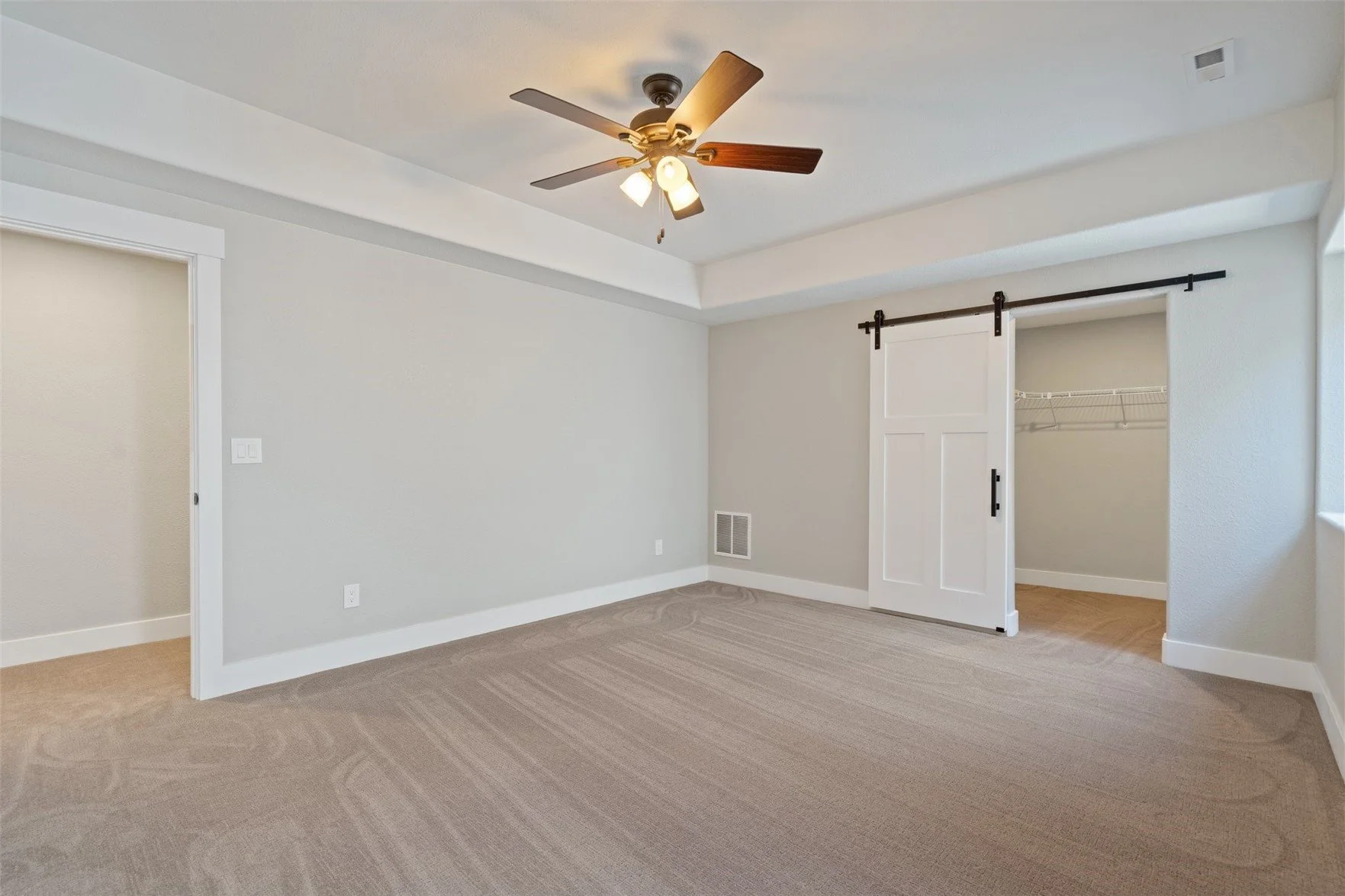 Empty bedroom with beige carpet, white walls, a ceiling fan with lights, a sliding barn door to a closet, and a vent on the wall.