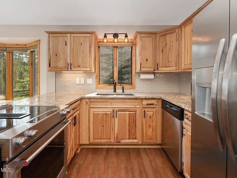 Condo kitchen with wooden cabinets, granite countertops, and stainless steel appliances including a refrigerator, oven, and dishwasher. A window above the sink shows trees outside.