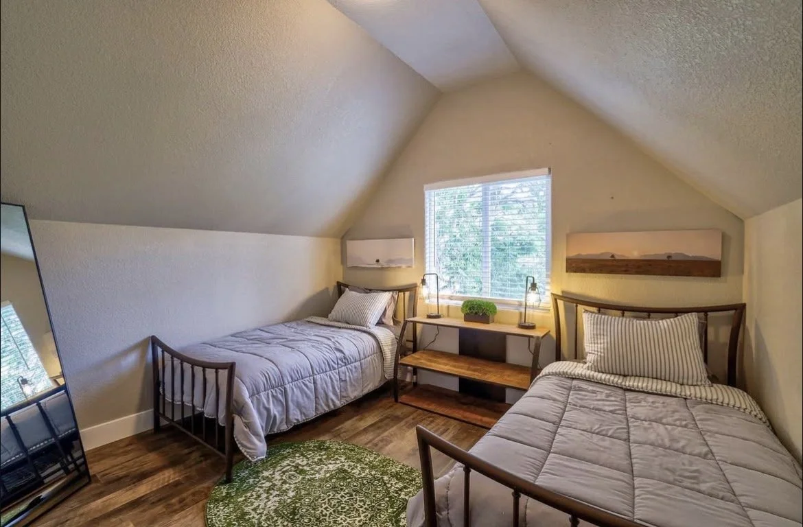 A cozy bedroom with two twin beds, a window with blinds, a small desk with lamps, a mirror leaning against the wall, and artwork on the walls.