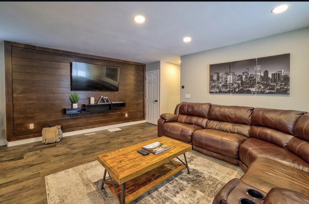 Living room with wood-paneled accent wall, mounted flat-screen TV, and decorative items. Brown leather sectional sofa, wooden coffee table, black and white cityscape art, and hardwood floors with area rug.