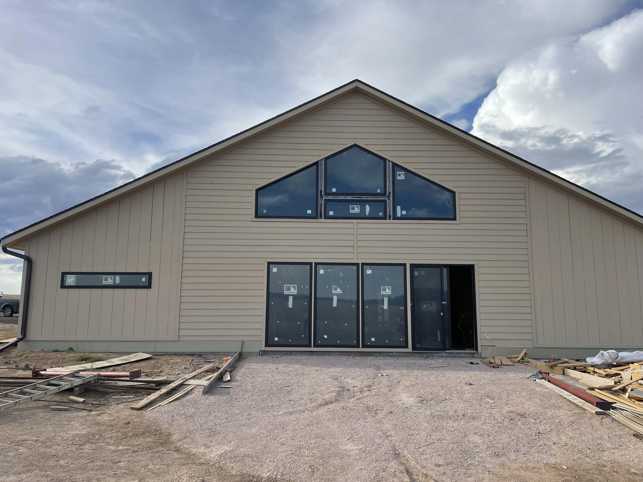 Construction site with a newly built house featuring beige siding, black framed windows, and a gravel ground with construction materials around.
