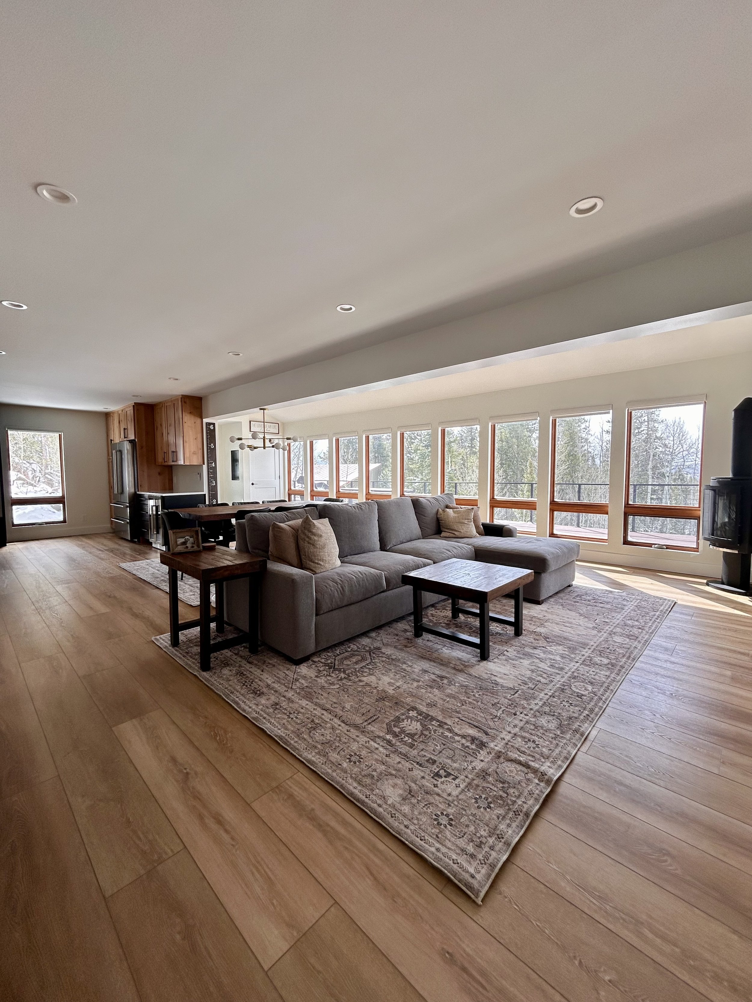 Open living room with large gray sectional sofa, wooden side tables, area rug, and large windows showing outdoor trees and snow.
