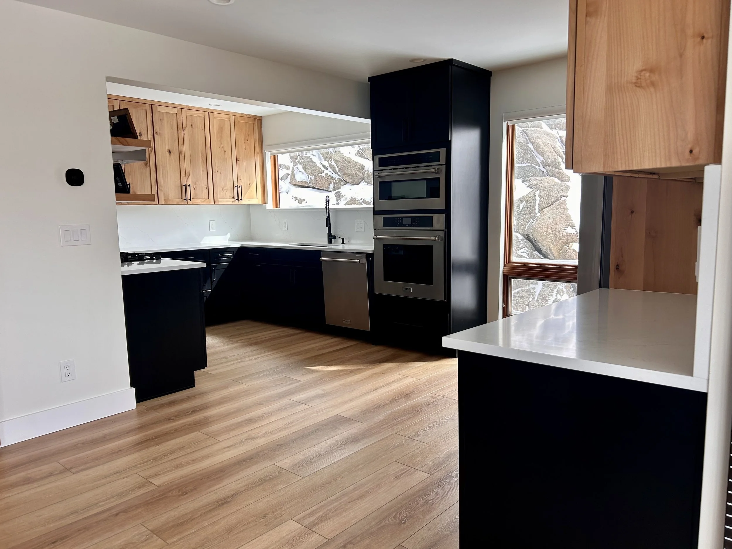 Modern mountain kitchen with light wooden cabinets, black lower cabinets, white countertops, and stainless steel appliances, including a double oven. There are large windows with a view of rock formations and snow outside.