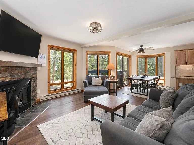 Living room condo with large windows, a gray sofa and armchair, a wooden coffee table, a fireplace with a flat-screen TV above, a dining table with chairs, and a kitchen area in the background.