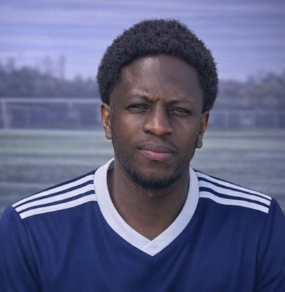 A man with short curly hair, wearing a blue sports jersey with white stripes on the shoulders, standing outdoors on a sports field with a cloudy sky in the background.