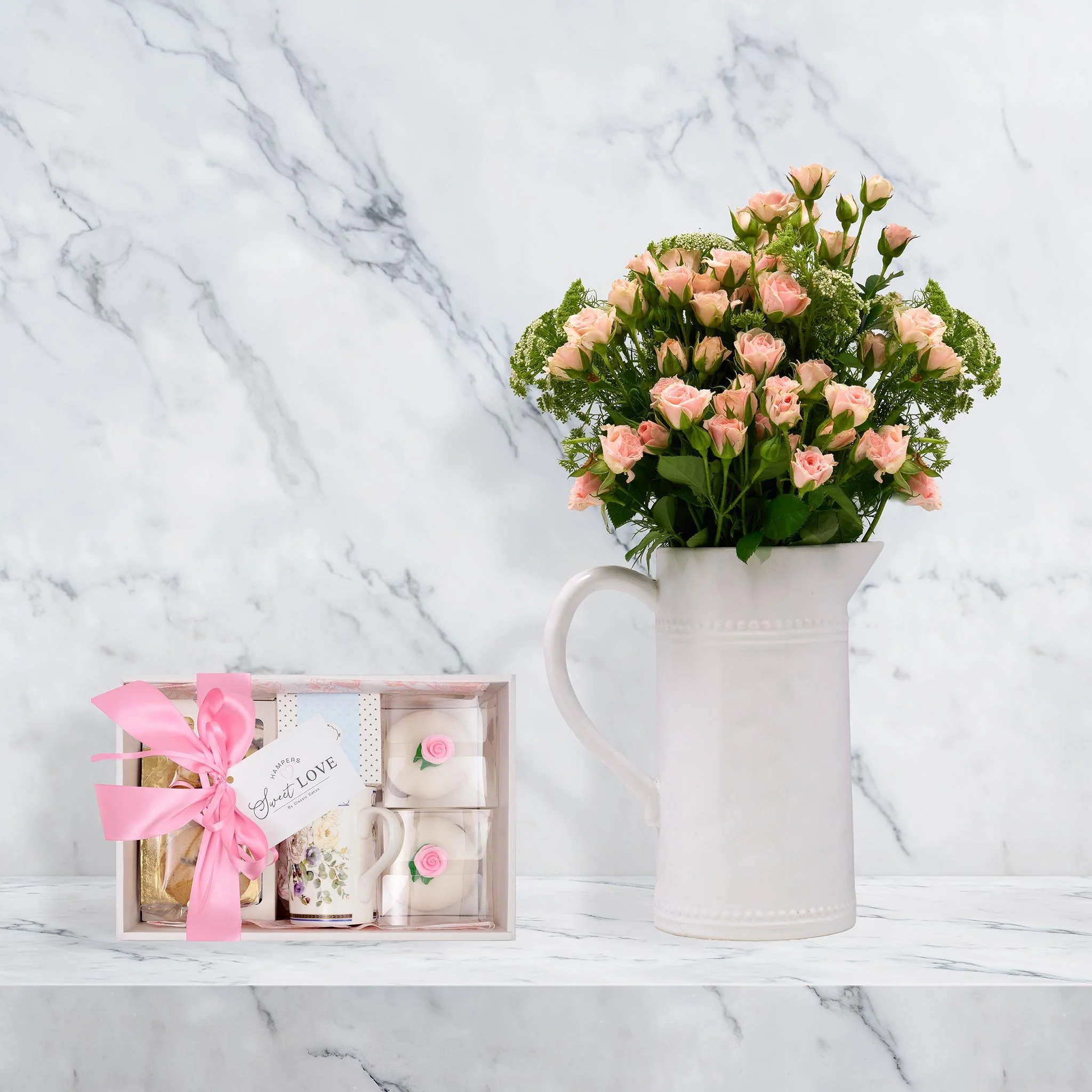 A large white ceramic pitcher with a handle, filled with a bouquet of pink roses and green foliage, placed on a white marble surface with a marble wall background. To the left, there is a gift box containing a mug with a floral design, two white cand