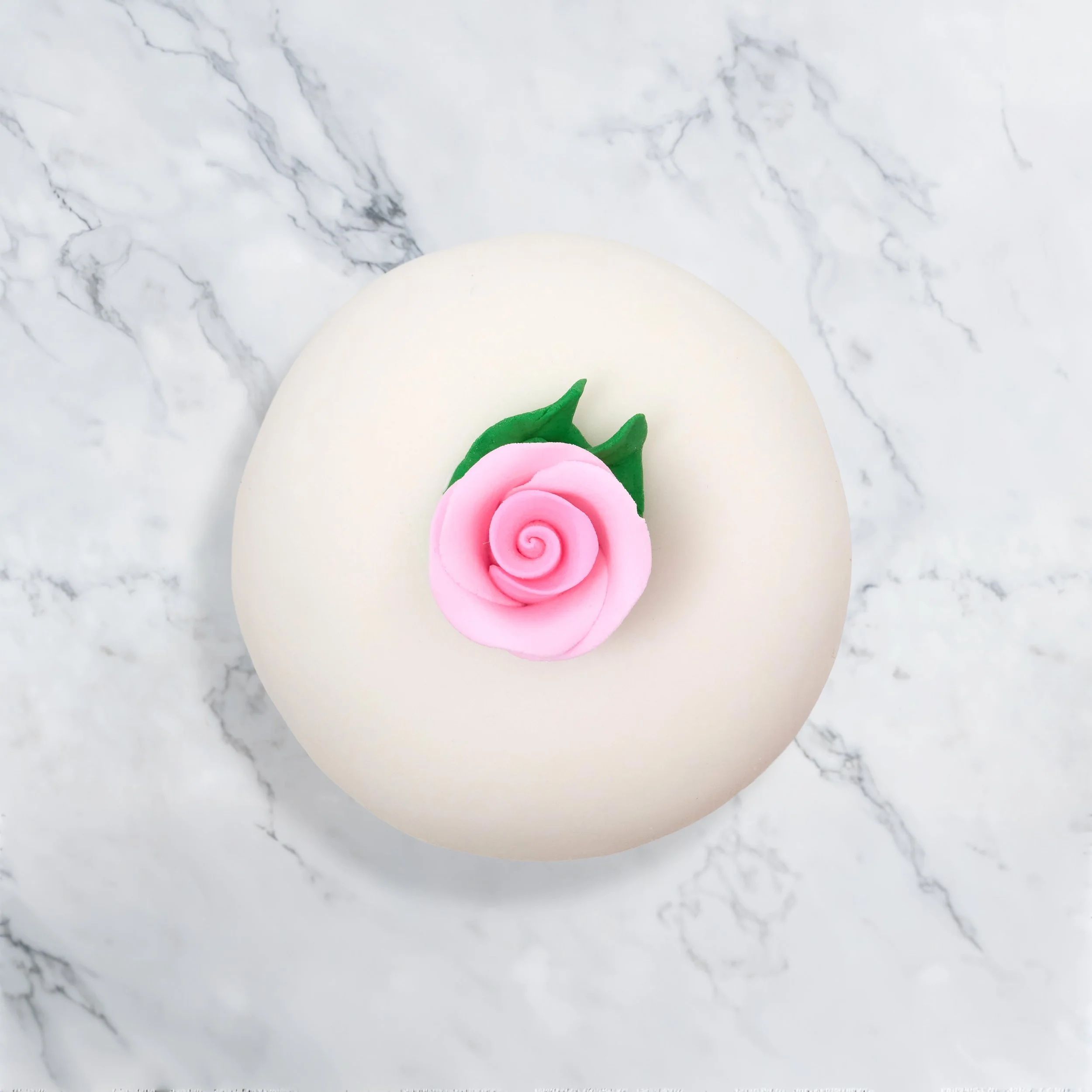 Round white cake with a pink fondant rose and green leaves on top, placed on a white marble surface.