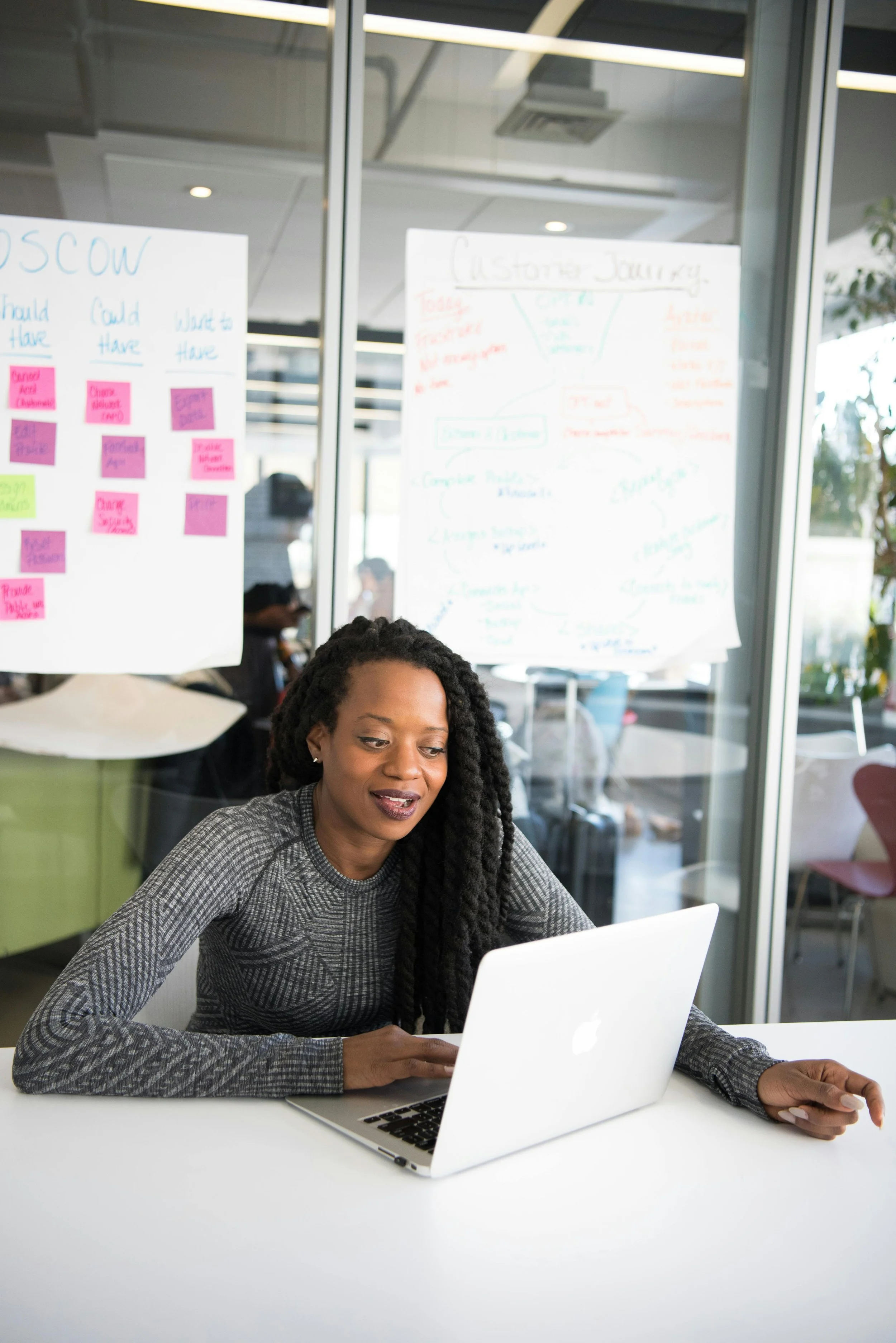 Woman with long braided hair working on a laptop in a modern office with whiteboards and sticky notes in the background.