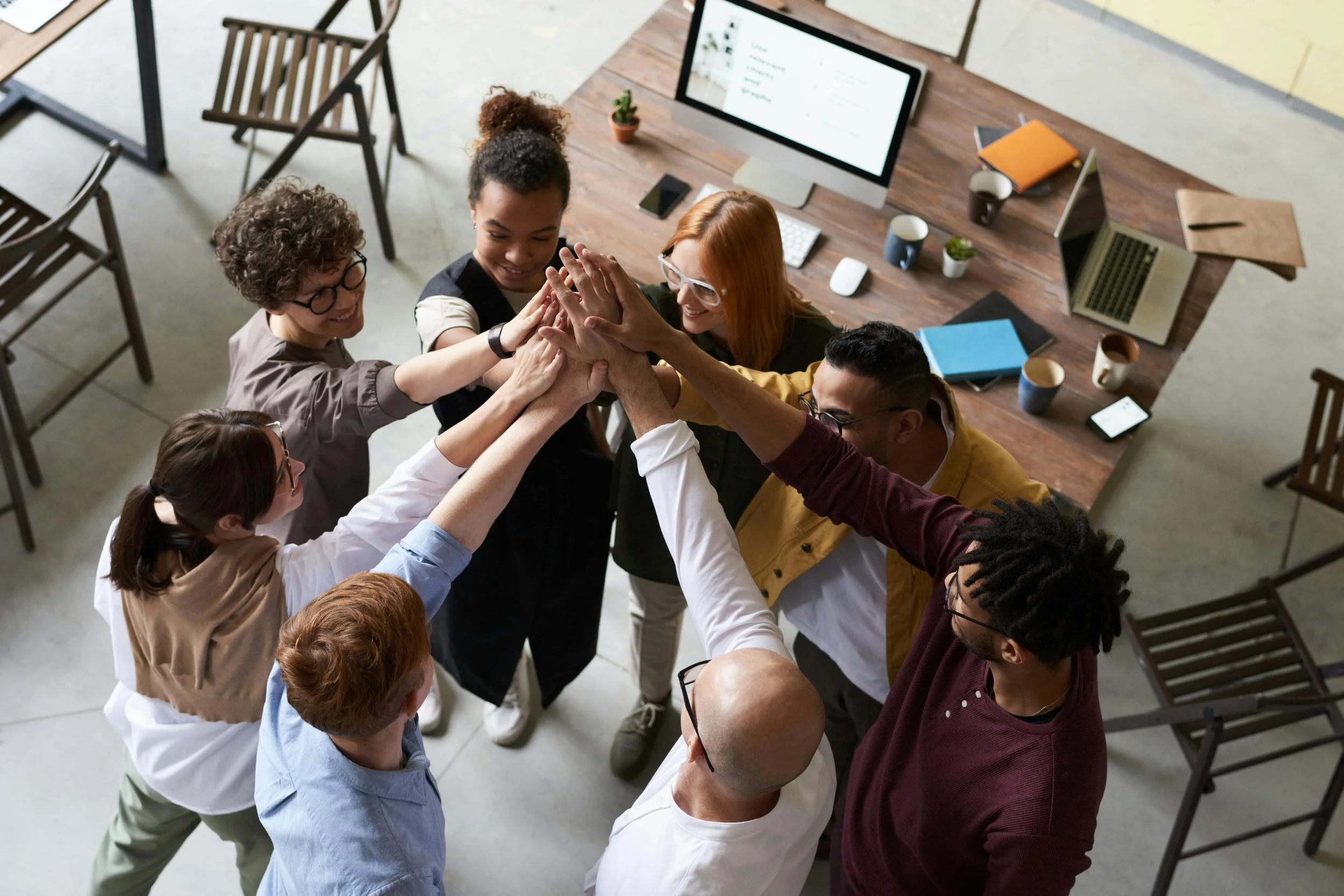 Team of diverse colleagues doing a group high five around a wooden office table with laptops and notepads.