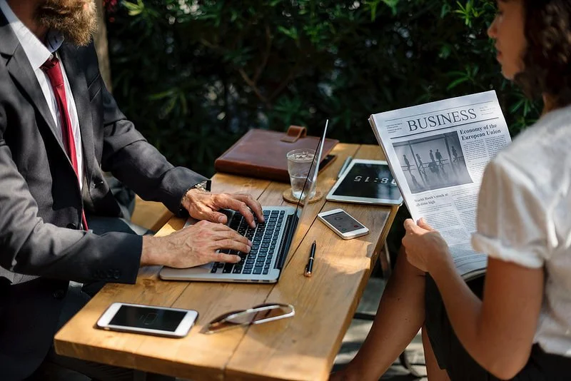 Two people sitting at an outdoor wooden table; one using a laptop, the other reading a business newspaper. Items on the table include a smartphone, tablet, pen, sunglasses, and a glass of water.