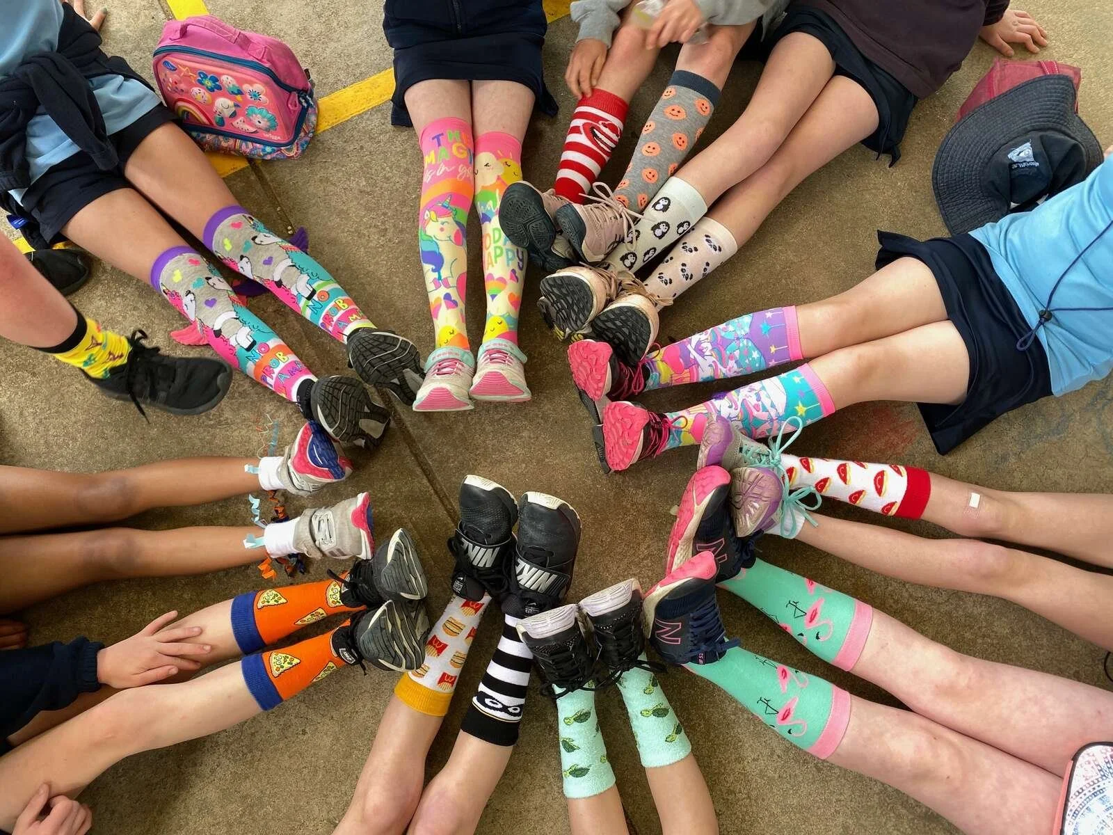 Children sitting in a circle with their legs extended inward, showcasing colorful and patterned socks and various sneakers, on a school gymnasium floor.