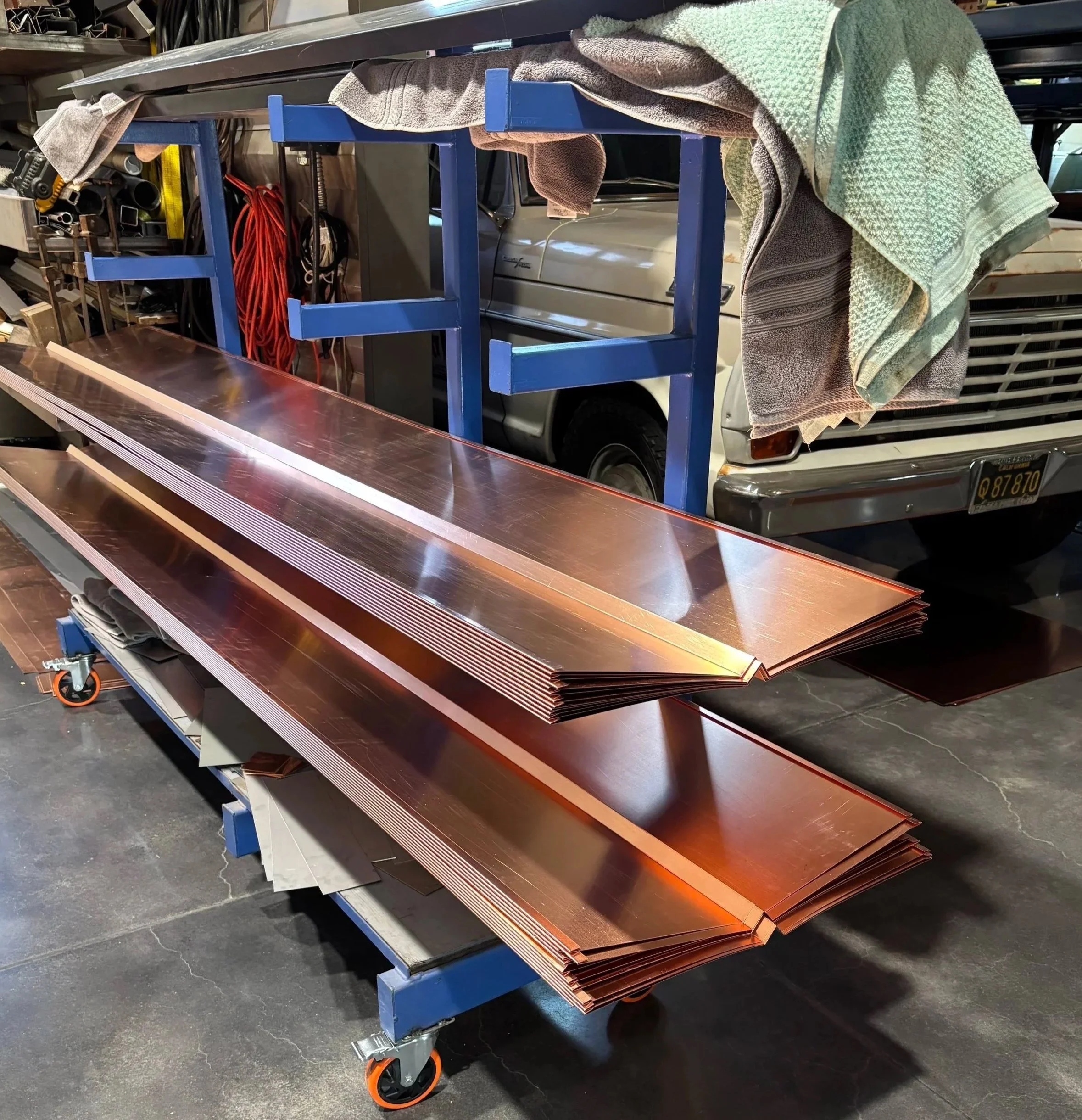 A blue cart with orange and black wheels holding a large sheet of copper-colored metal and several towels draped over the top, situated in a workshop or garage with tools and equipment in the background.